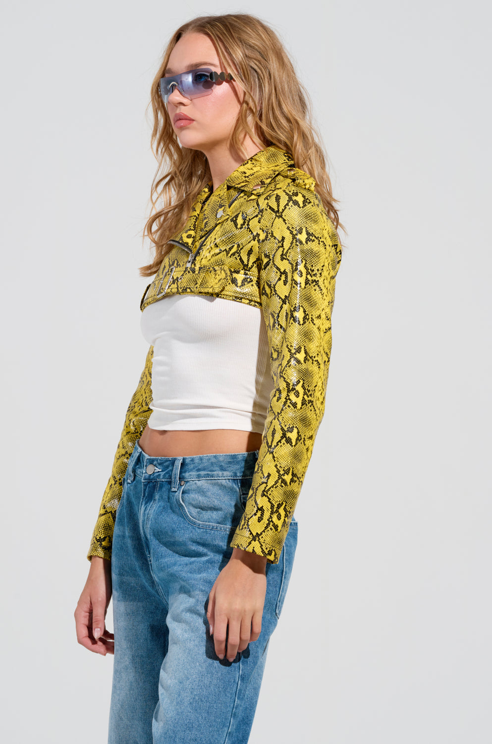 A person wearing blue sunglasses and a yellow faux snakeskin SYLVETTE ULTRA CROP MOTO over a white top with blue jeans stands against a plain background.
