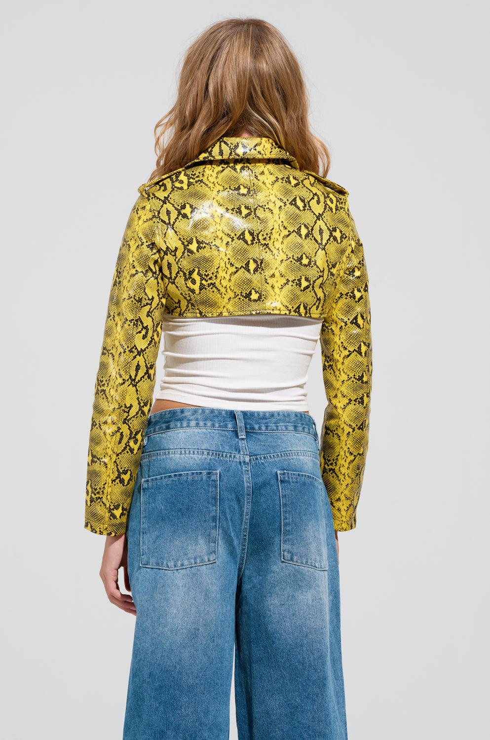 A person with long hair stands with their back to the camera, showcasing the SYLVETTE ULTRA CROP MOTO jacket in vibrant yellow and black faux snakeskin. They’ve paired it beautifully with a white top and blue jeans, creating a striking ensemble that accentuates the jacket’s adjustable waist belt.