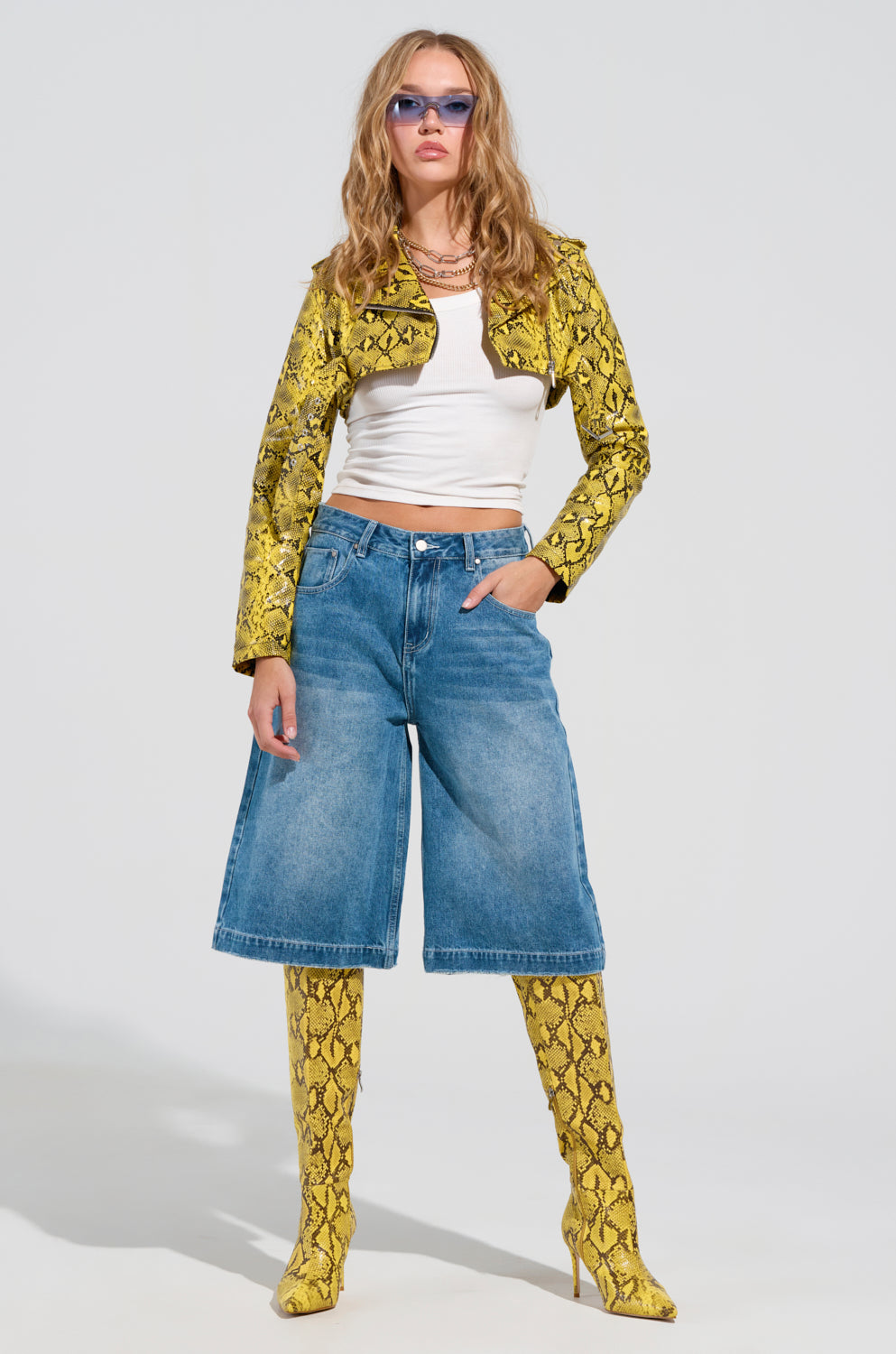 A person poses confidently in a SYLVETTE ULTRA CROP MOTO jacket, featuring an adjustable waist belt in striking yellow snakeskin. They pair it with matching knee-high boots, blue denim culottes, a white top, and blue-tinted sunglasses. Standing against a plain light background, they exude a trendy, fashion-forward vibe.