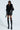 A person confidently stands against a plain background wearing the "ON THE GO FUR TRIM MOTO IN BLACK," complemented by matching leather boots, sunglasses, knee-high stockings, and a short skirt. Their bold and stylish look is accentuated by the faux leather jacket's fur-embellished collar.