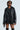 Dressed in the ON THE GO FUR TRIM MOTO IN BLACK, a person exudes style with a faux leather jacket featuring a quilted interior and pairs it effortlessly with a pinstriped dress. Star-shaped earrings dangle beneath their sunglasses as they pose confidently against a light background.