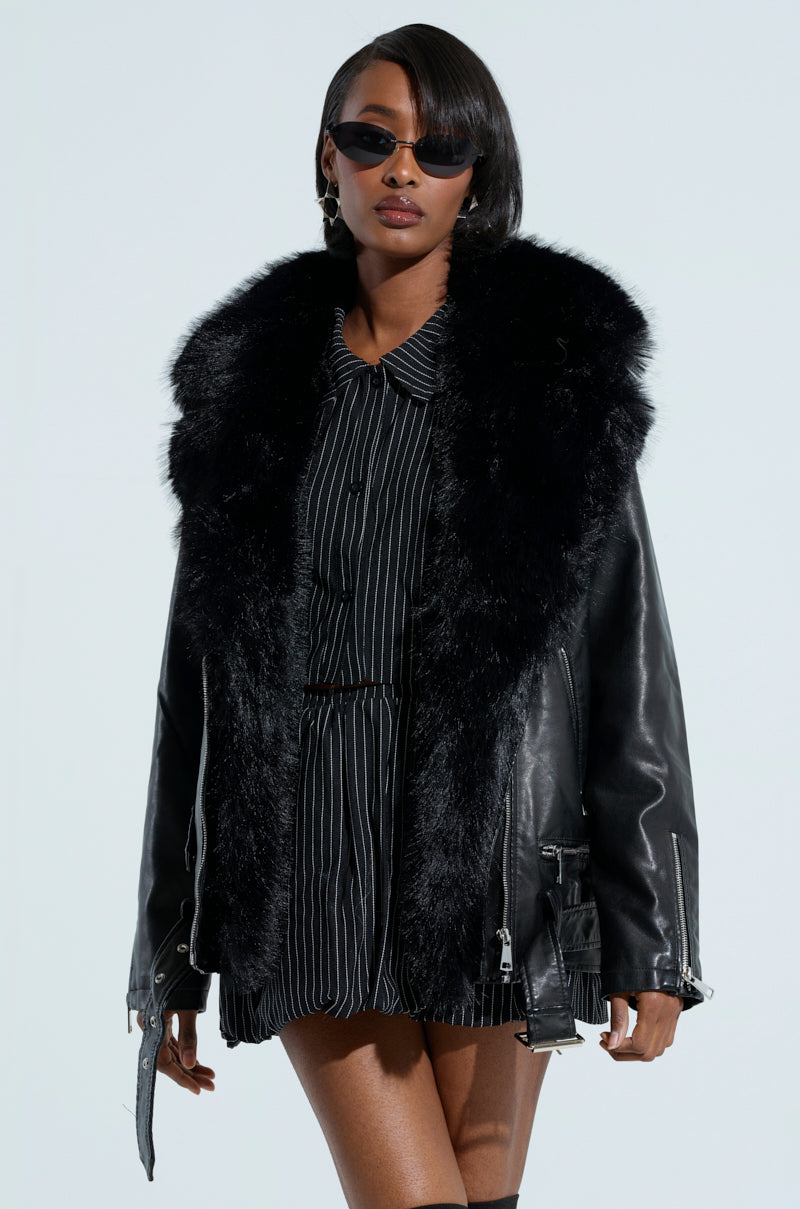 A woman wearing sunglasses and the ON THE GO FUR TRIM MOTO IN BLACK jacket over a pinstriped dress. Her hair is styled in a bob, and she stands against a light gray background.