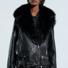 A person wearing the ON THE GO FUR TRIM MOTO IN BLACK, showcasing its chic faux leather design with a large faux fur collar, stylish zippers, and belt buckle detail. They're also sporting black sunglasses and a pinstriped outfit underneath, set against a plain, light-colored background.