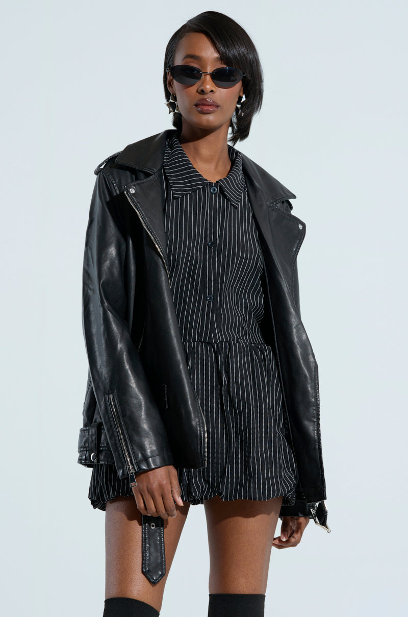 A person wearing the ON THE GO FUR TRIM MOTO IN BLACK over a striped dress stands confidently. They have sunglasses on and knee-high socks, exuding a cool and stylish vibe against a minimalistic background.