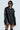 A person wearing the ON THE GO FUR TRIM MOTO IN BLACK over a striped dress stands confidently. They have sunglasses on and knee-high socks, exuding a cool and stylish vibe against a minimalistic background.