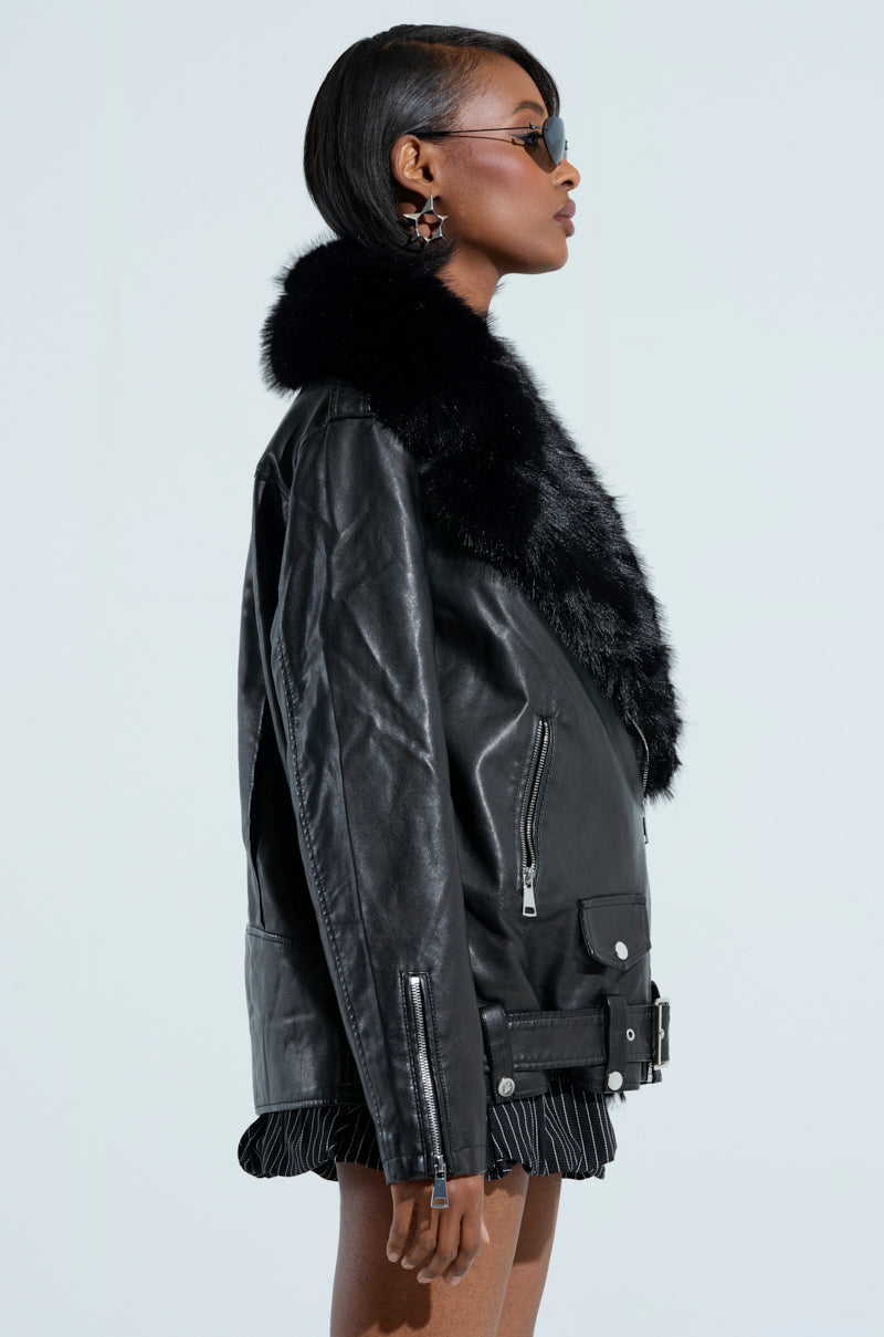 A person in the "ON THE GO FUR TRIM MOTO IN BLACK" jacket with sunglasses stands in profile against a light background. The black leather jacket boasts a quilted interior lining and faux fur collar, flawlessly complementing their short hair and striped skirt.