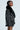 A person in the "ON THE GO FUR TRIM MOTO IN BLACK" jacket with sunglasses stands in profile against a light background. The black leather jacket boasts a quilted interior lining and faux fur collar, flawlessly complementing their short hair and striped skirt.