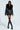A person wearing the "ON THE GO FUR TRIM MOTO IN BLACK" jacket, featuring a quilted interior lining and fur collar, paired with a black mini skirt and knee-high lace-up boots. They have sunglasses and thigh-high stockings, standing against a light background.