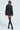A person poses confidently wearing an ON THE GO FUR TRIM MOTO IN BLACK jacket over a black pinstriped dress, paired with mid-calf lace-up boots and dark sunglasses. Their stylish, edgy appearance is completed with long earrings and stockings.