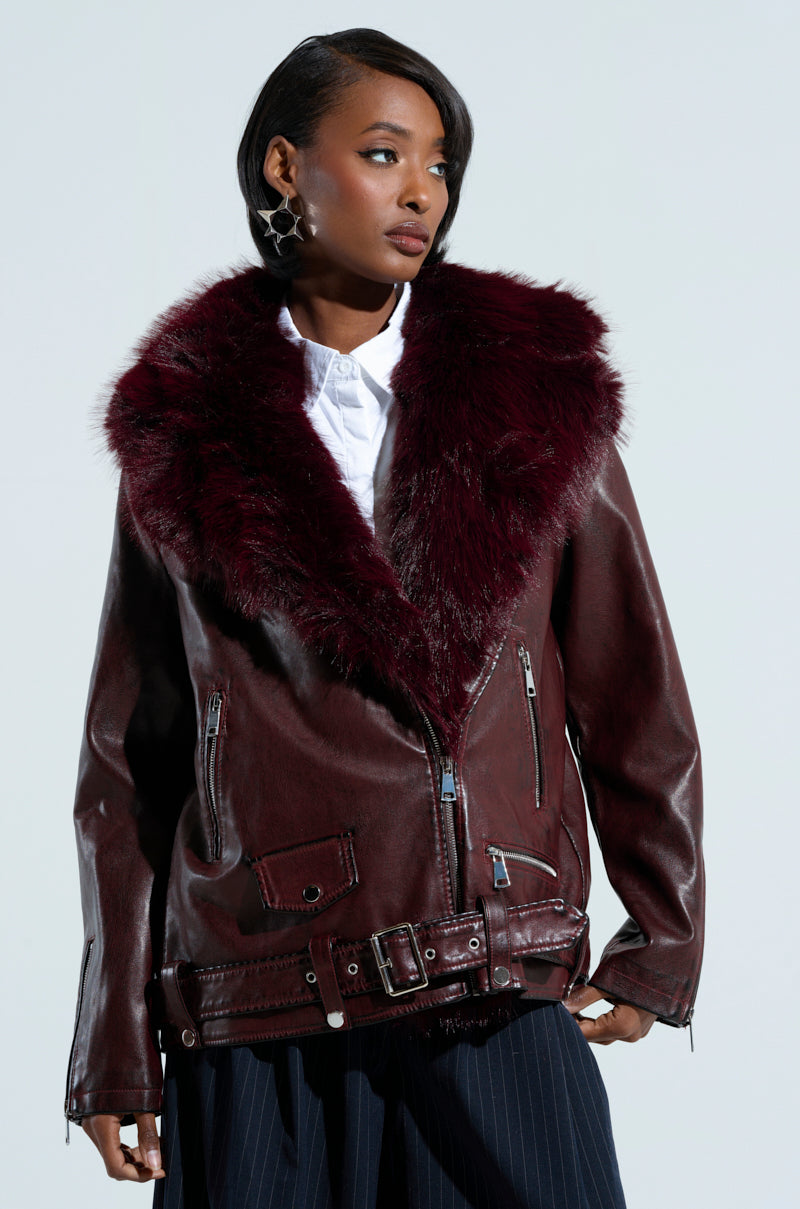 A person confidently dons the ON THE GO FUR TRIM MOTO IN BURGUNDY, featuring a deep burgundy hue with a plush faux fur collar. This faux leather jacket is adorned with multiple zippers and a waist belt. Underneath, they wear a white shirt, and their short, styled hair complements the neutral gray background.