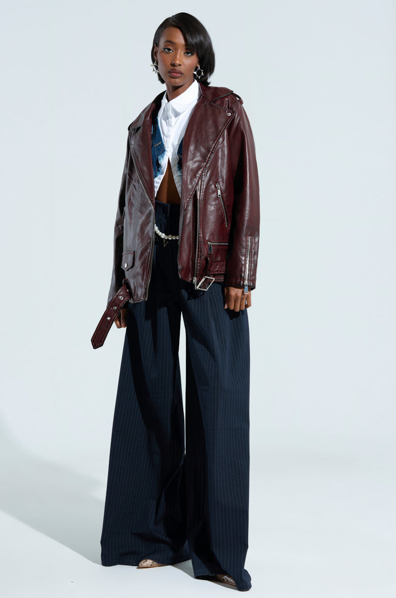 A person dressed in a white shirt, the "On The Go Fur Trim Moto in Burgundy," dark wide-leg trousers, and beige shoes is standing against a light background. They have short hair and are holding a small object in one hand.