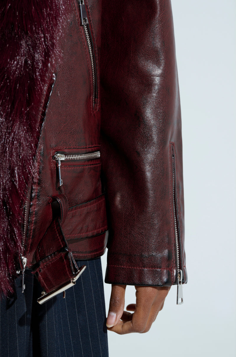 In the spotlight is a close-up of an individual modeling the ON THE GO FUR TRIM MOTO IN BURGUNDY jacket, featuring dark brown faux leather with silver zippers and maroon faux fur trim, paired with pinstriped black pants. The emphasis is on the sleeve and detailed accents of the jacket.