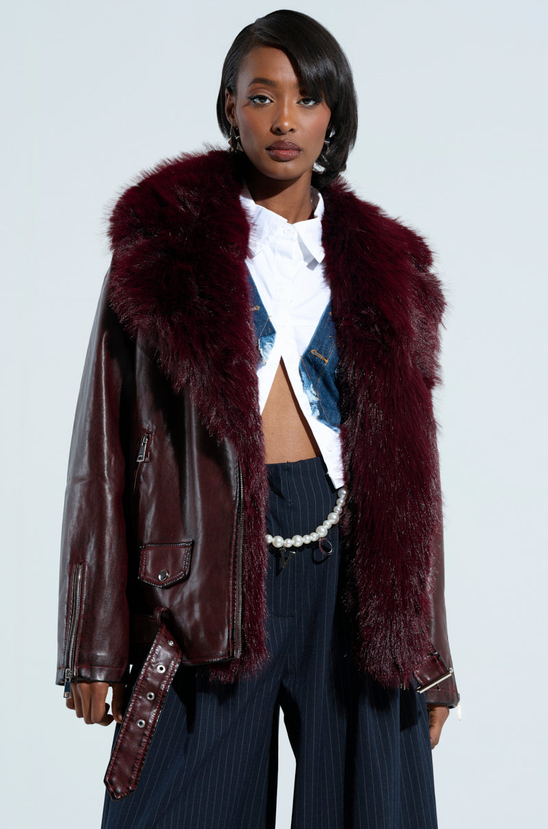 A person wearing the "ON THE GO FUR TRIM MOTO IN BURGUNDY," a jacket crafted from burgundy faux leather and featuring a prominent faux fur collar, paired with a white shirt and pinstriped pants. The ensemble is completed with a chain belt, set against a plain, light-hued backdrop.