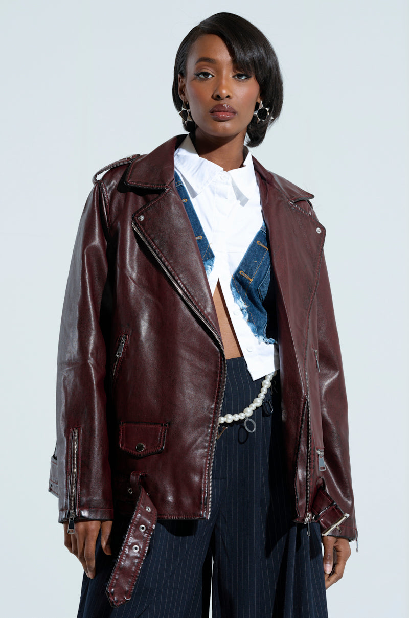 Dressed confidently, an individual sports the ON THE GO FUR TRIM MOTO in burgundy over a denim vest and white shirt, paired with pinstripe pants. Their side-parted hair complements the chic style as they pose against a plain background.