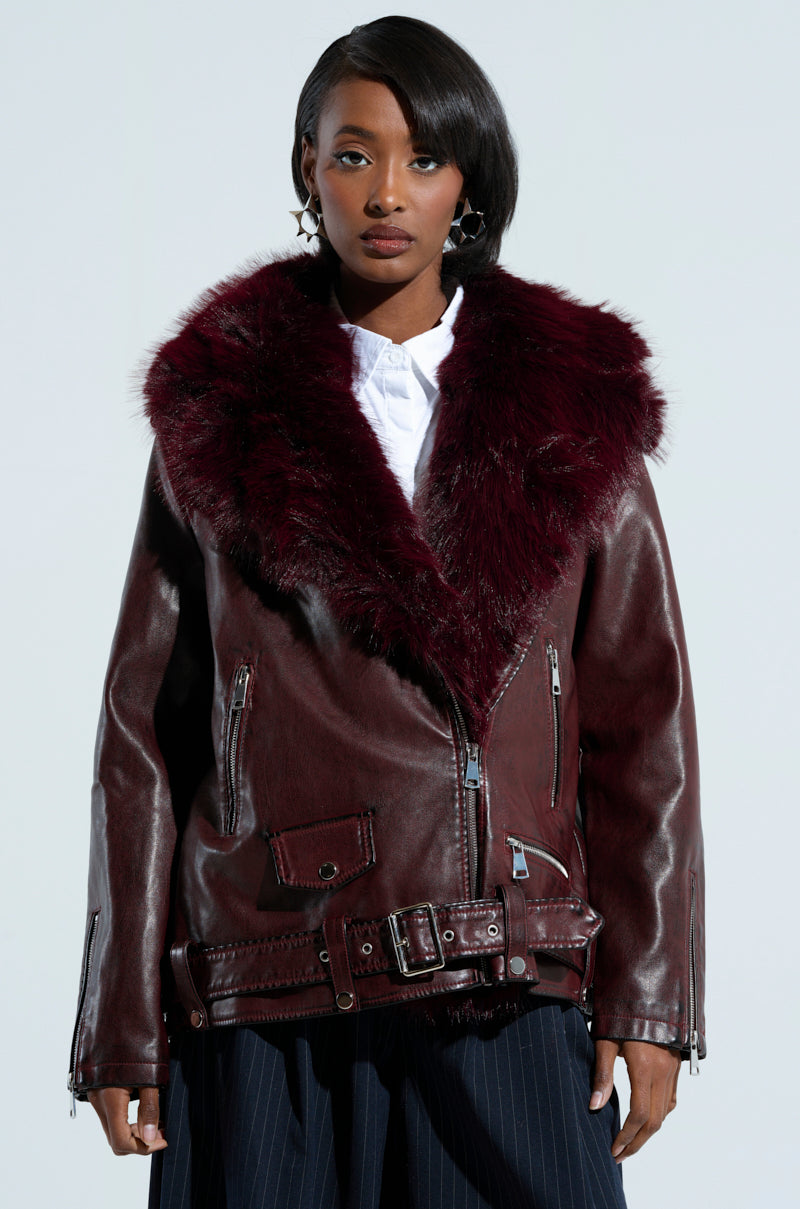 A person is wearing the fashionable "ON THE GO FUR TRIM MOTO IN BURGUNDY," a dark burgundy faux leather jacket with a large faux fur collar, multiple zippers, and a belt. They have shoulder-length hair and are standing against a plain background.