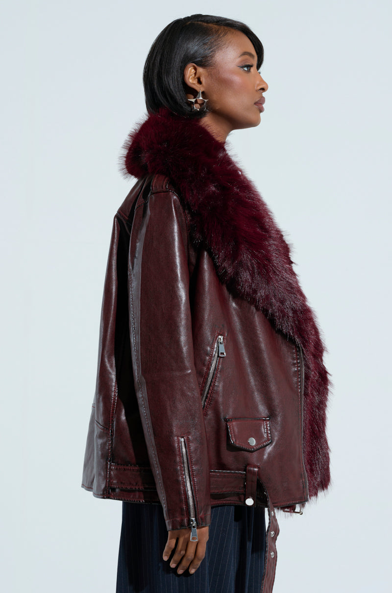 A person is shown from the side, confidently posing in the "ON THE GO FUR TRIM MOTO IN BURGUNDY" jacket. The piece features a rich burgundy faux leather with a large faux fur collar. They have short hair and stand against a plain background, exuding effortless style.