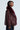 A person is shown from the side, confidently posing in the "ON THE GO FUR TRIM MOTO IN BURGUNDY" jacket. The piece features a rich burgundy faux leather with a large faux fur collar. They have short hair and stand against a plain background, exuding effortless style.