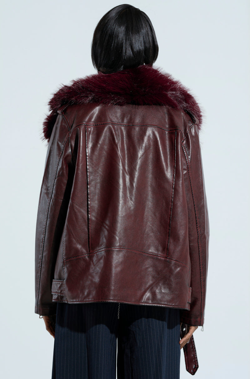 A person stands facing away, wearing the 'ON THE GO FUR TRIM MOTO IN BURGUNDY,' a dark brown jacket featuring a dark red faux fur collar. The jacket is paired with dark pants against a plain light background.