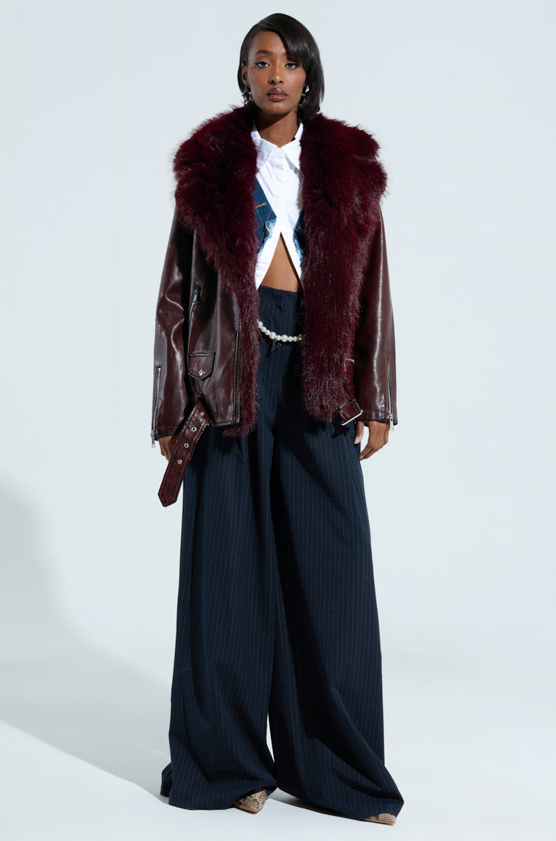 A person stands confidently on a neutral background, showcasing a stylish outfit that highlights the ON THE GO FUR TRIM MOTO IN BURGUNDY, paired with a cropped white shirt and wide-leg pinstripe pants.