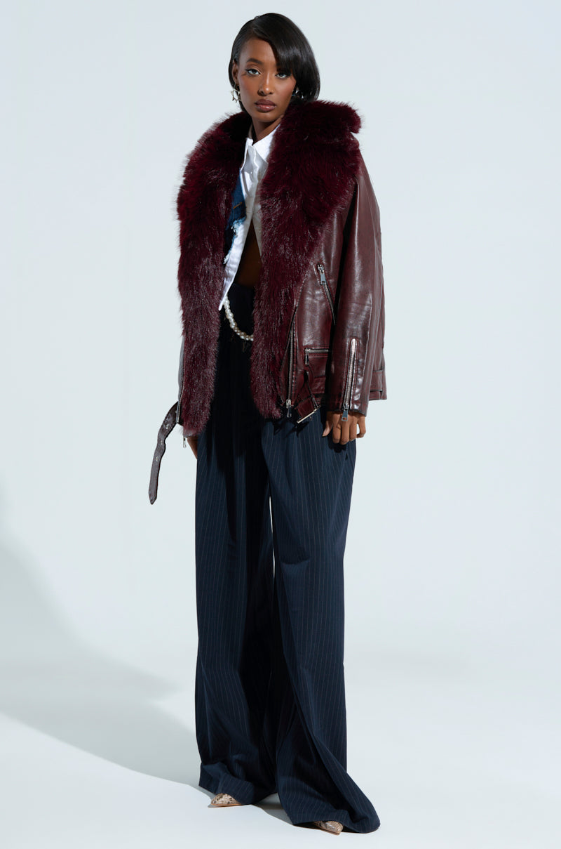 A person showcases a chic ensemble with the ON THE GO FUR TRIM MOTO jacket in burgundy, featuring a large faux fur collar and black faux leather design. This is complemented by wide-leg pinstriped pants and a partially unbuttoned white shirt. The setting is plain and well-lit.