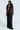 A person showcases a chic ensemble with the ON THE GO FUR TRIM MOTO jacket in burgundy, featuring a large faux fur collar and black faux leather design. This is complemented by wide-leg pinstriped pants and a partially unbuttoned white shirt. The setting is plain and well-lit.