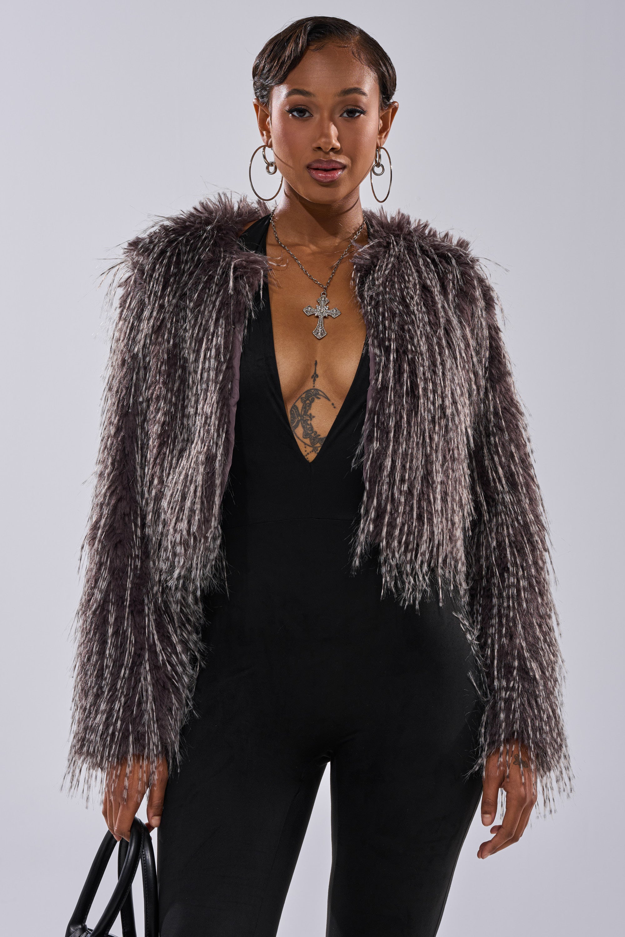 A woman wears the BIRDS OF A FEATHER SHAGGY FAUX FUR jacket over a black deep V-neck jumpsuit, accessorized with layered necklaces, hoop earrings, and a black handbag, standing against a plain background.