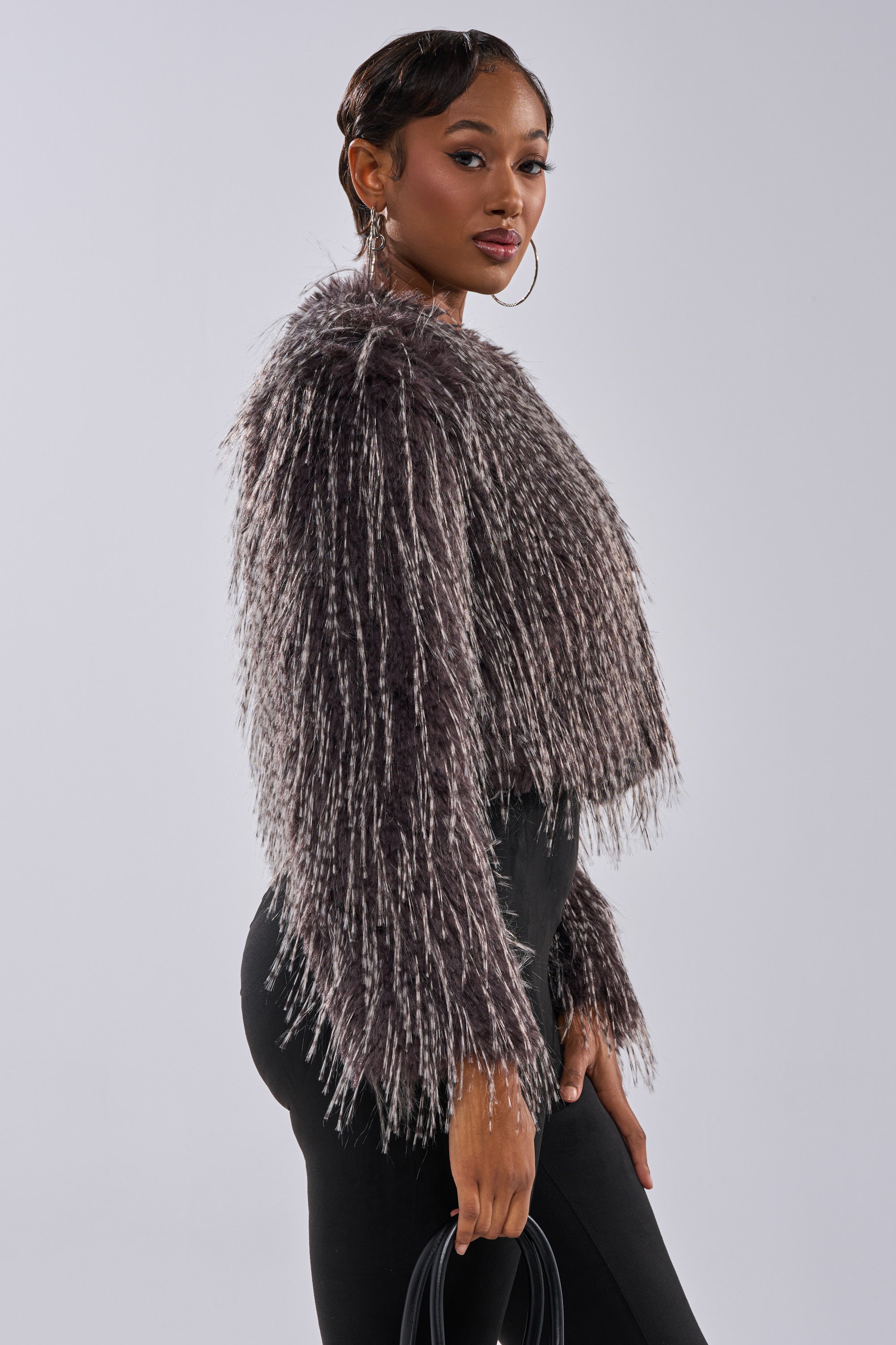 A woman poses against a plain background wearing the BIRDS OF A FEATHER SHAGGY FAUX FUR jacket, styled with hoop earrings, black pants, and a black handbag in hand.