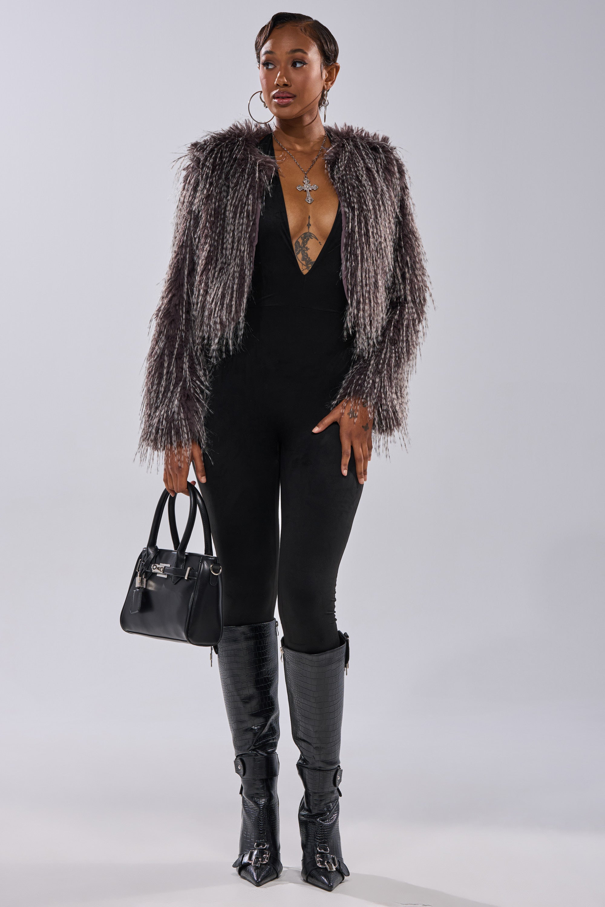 A stylish woman stands confidently in the BIRDS OF A FEATHER SHAGGY FAUX FUR jacket over a black plunging jumpsuit, paired with black knee-high boots and a black handbag, against a plain light background.