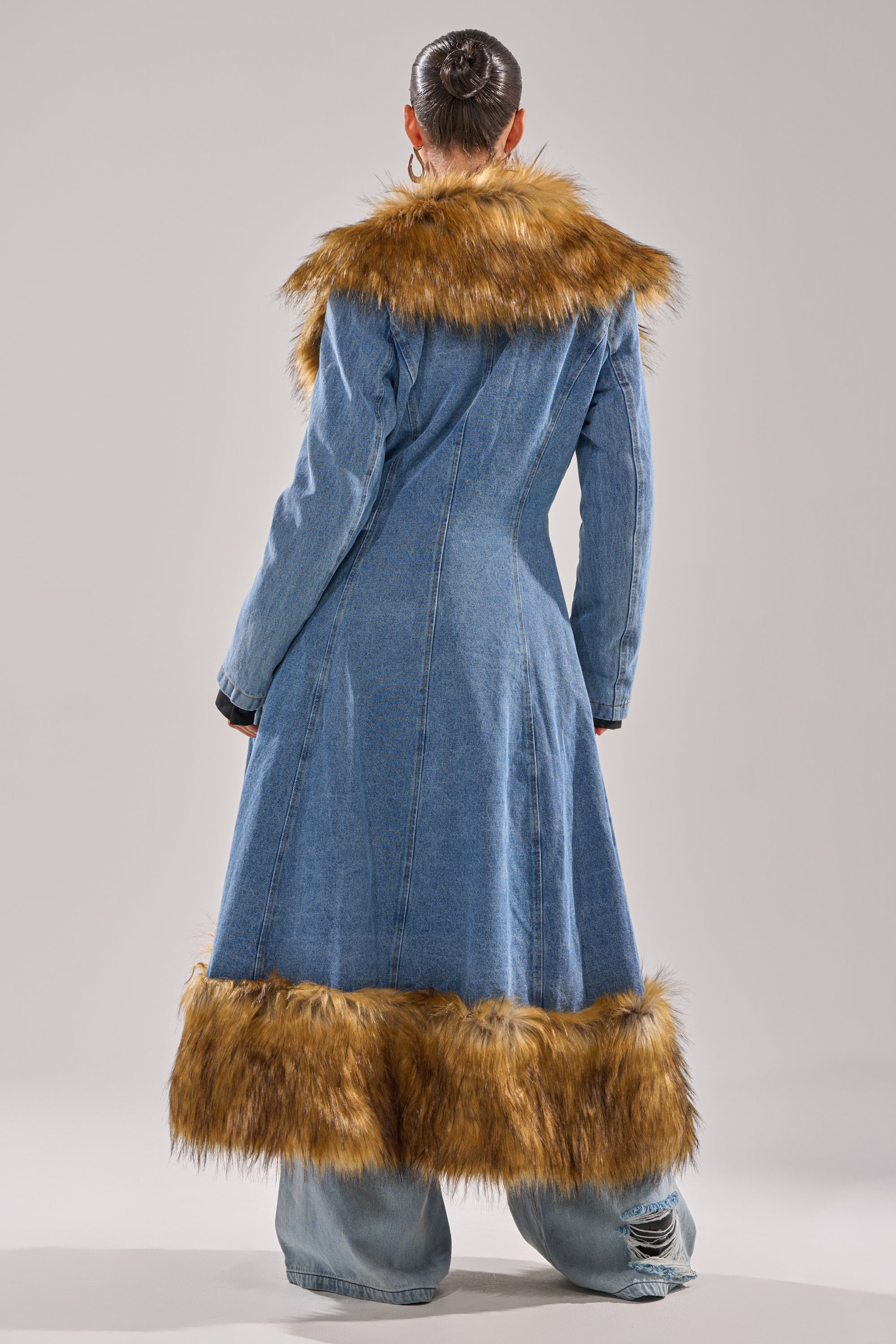 A person stands facing away in the VIVIAN FUR TRIM TRENCH IN DENIM, featuring brown faux fur at the collar and hem, styled with wide-leg distressed jeans and a sleek bun against a plain, light background.