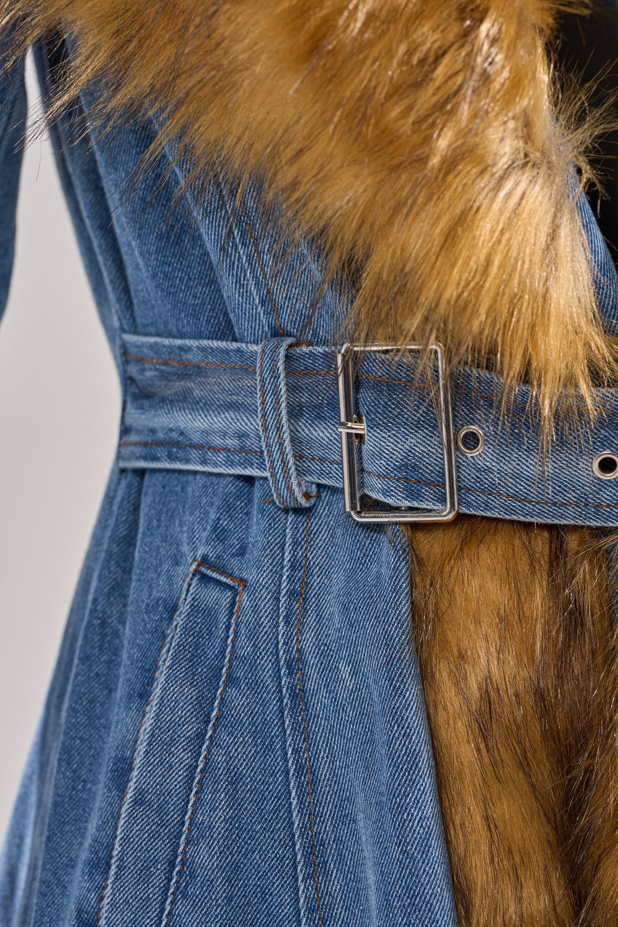 Close-up of the VIVIAN FUR TRIM TRENCH IN DENIM—blue denim coat with a matching belt and buckle, showcasing a striking brown faux fur collar and clearly visible fabric and fur textures.