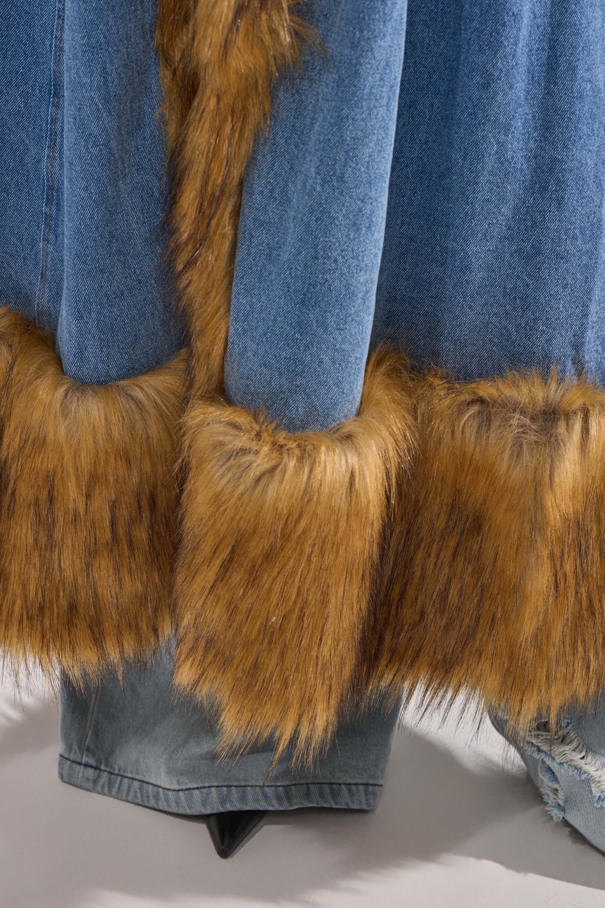 Close-up of the VIVIAN FUR TRIM TRENCH IN DENIM featuring brown faux fur on cuffs and hem, layered over denim pants. The soft fur detail adds a stylish contrast to the smooth blue denim.