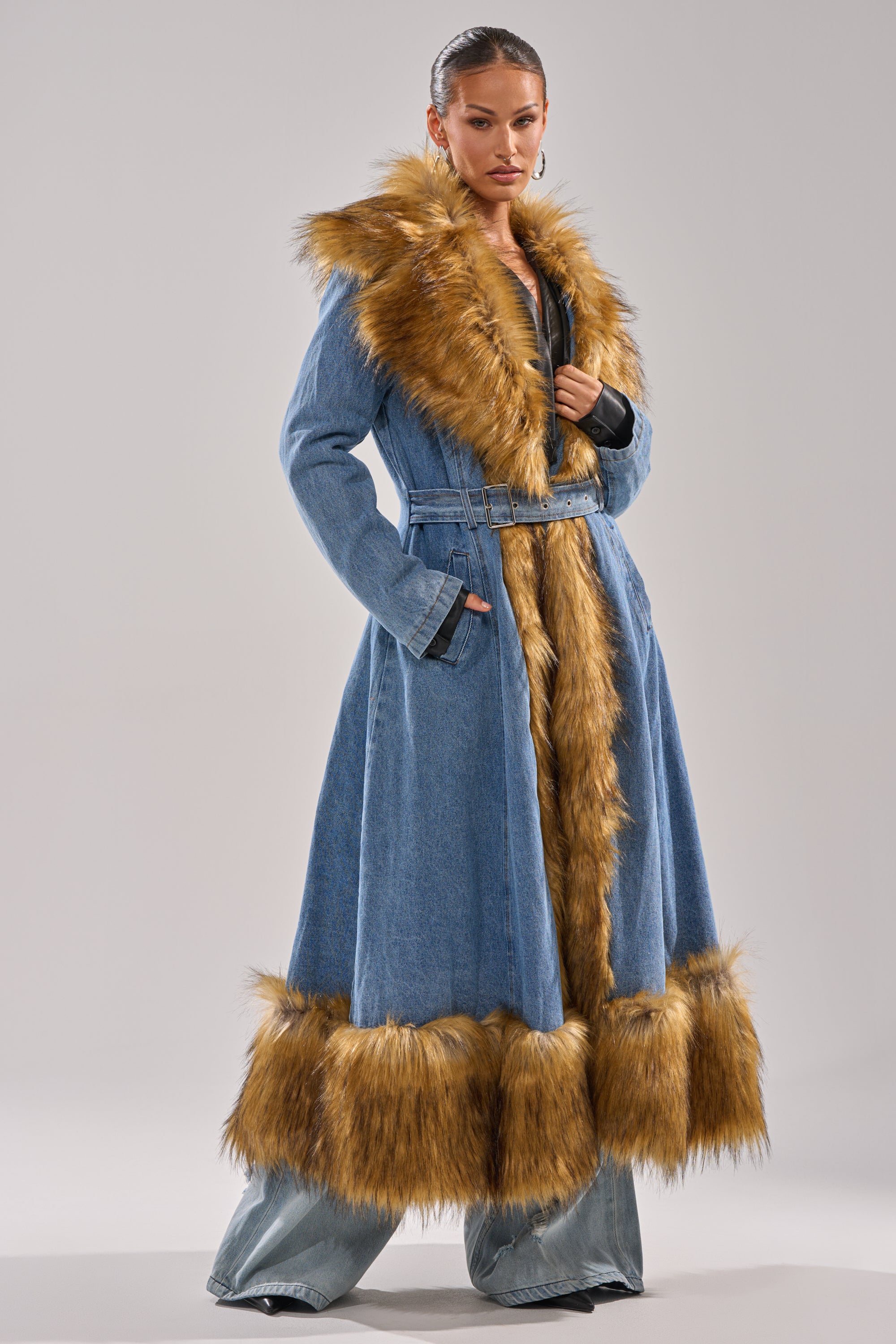 A woman poses confidently in the VIVIAN FUR TRIM TRENCH IN DENIM, a longline denim coat with wide golden brown faux fur trim on the collar, front, cuffs, and hem, standing against a plain light background.