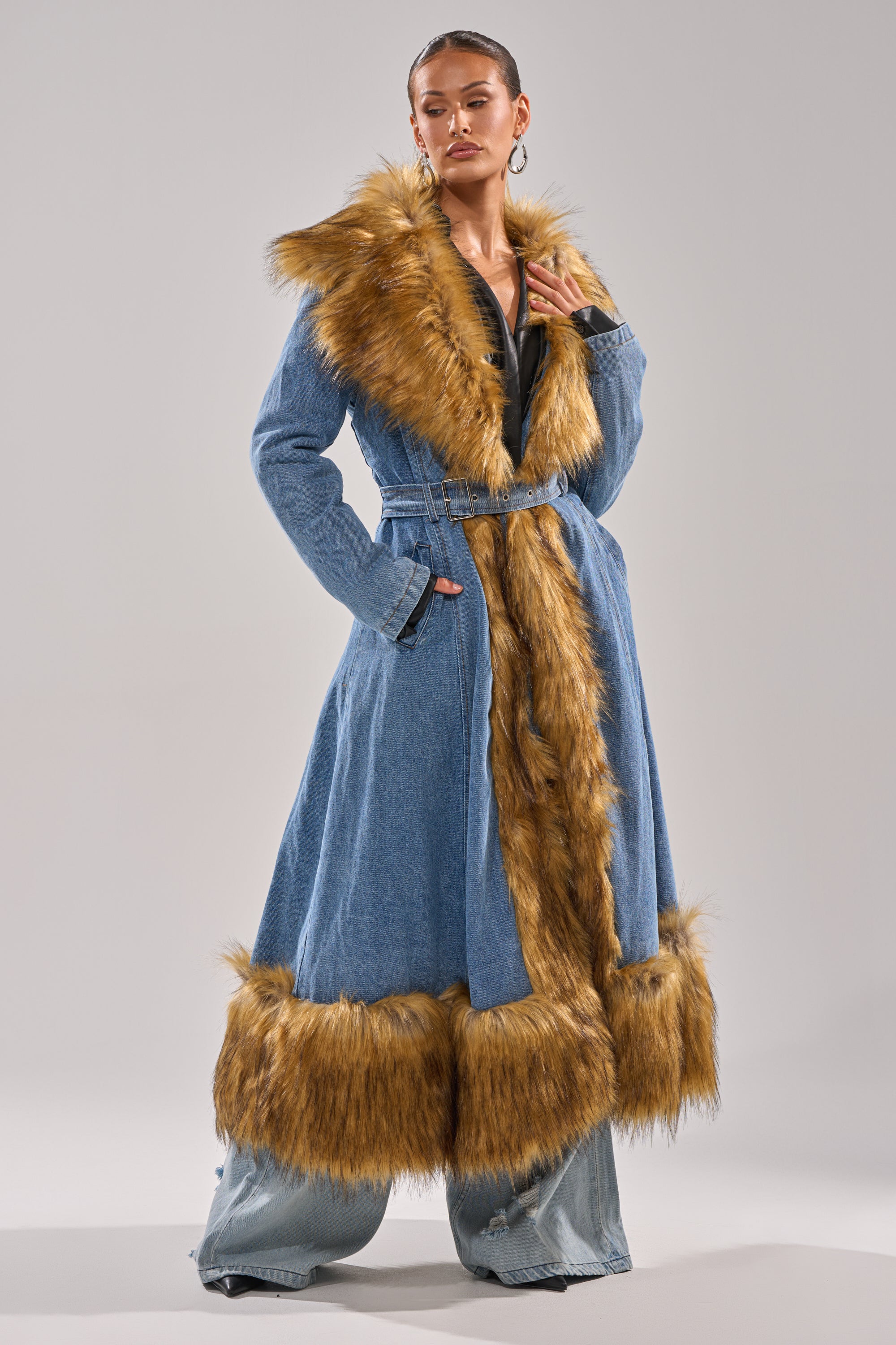 A woman poses against a neutral background wearing the VIVIAN FUR TRIM TRENCH IN DENIM, featuring a longline silhouette with wide brown faux fur trim, belted at the waist and styled with wide-leg denim pants.