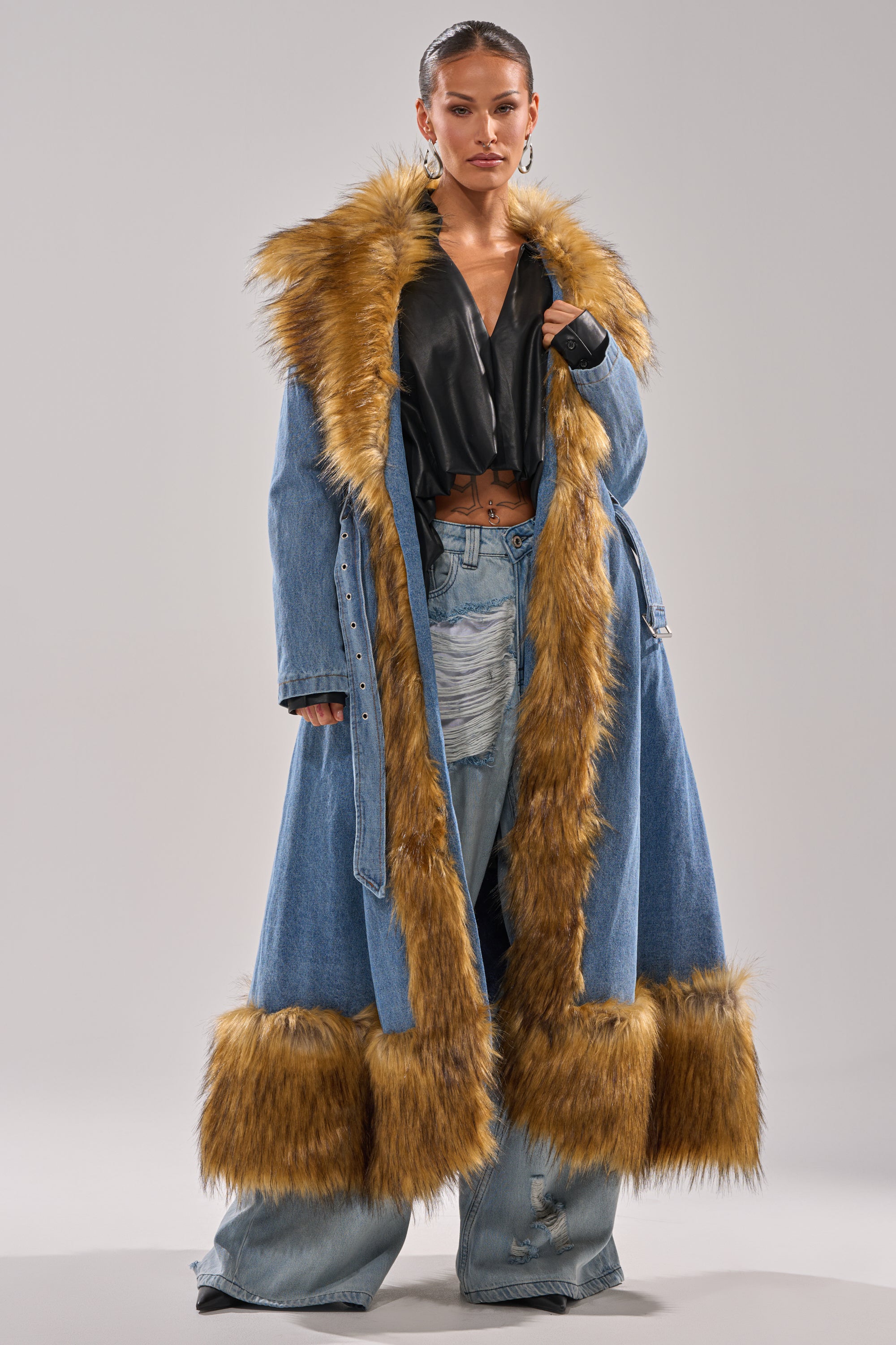 A woman exudes confidence in the VIVIAN FUR TRIM TRENCH IN DENIM, styled with a black button-up shirt, wide-legged distressed jeans, bold hoop earrings, and her hair pulled back.
