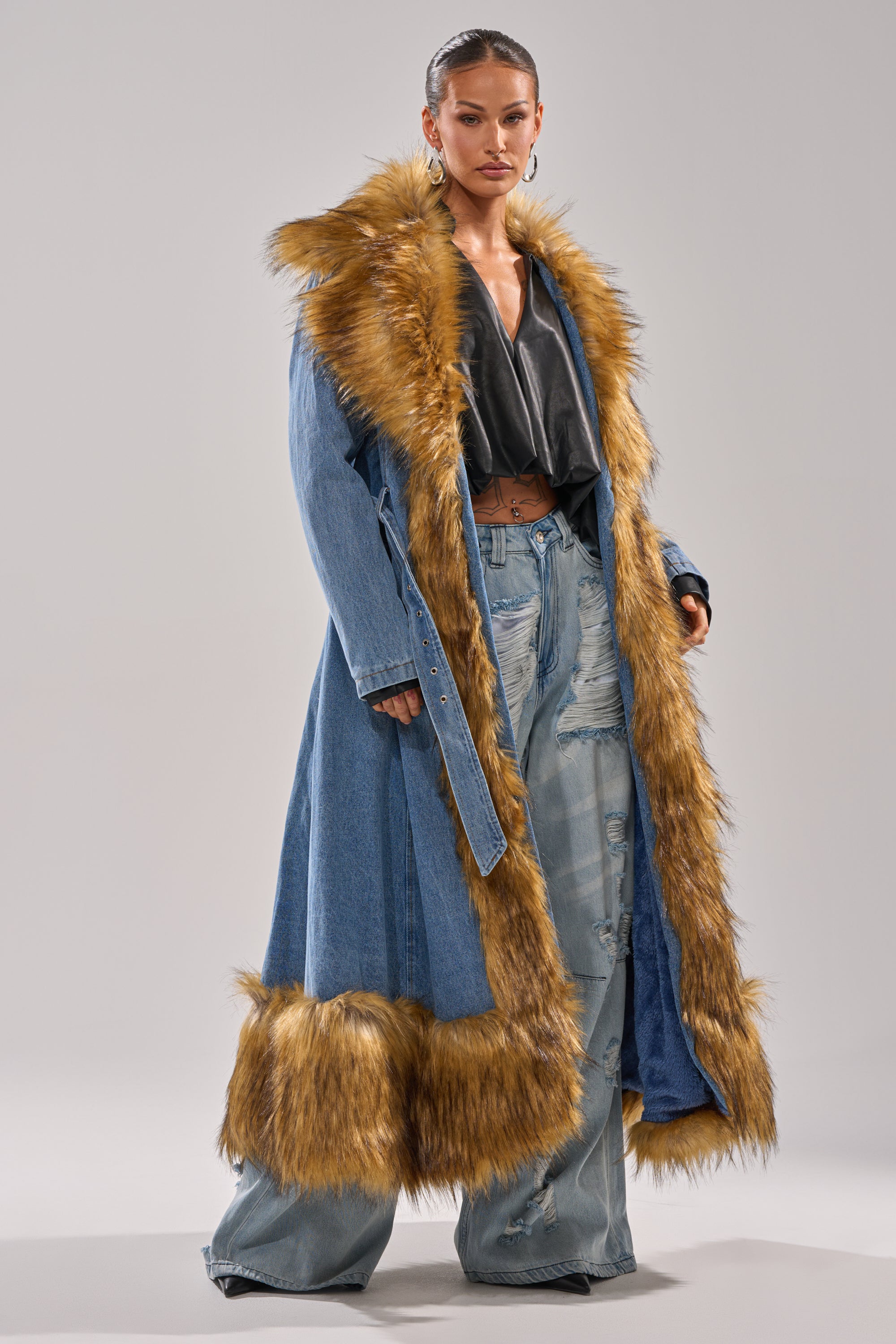 A woman confidently poses against a plain background in the VIVIAN FUR TRIM TRENCH IN DENIM, featuring brown faux fur trim, paired with a black top and wide-leg ripped jeans.