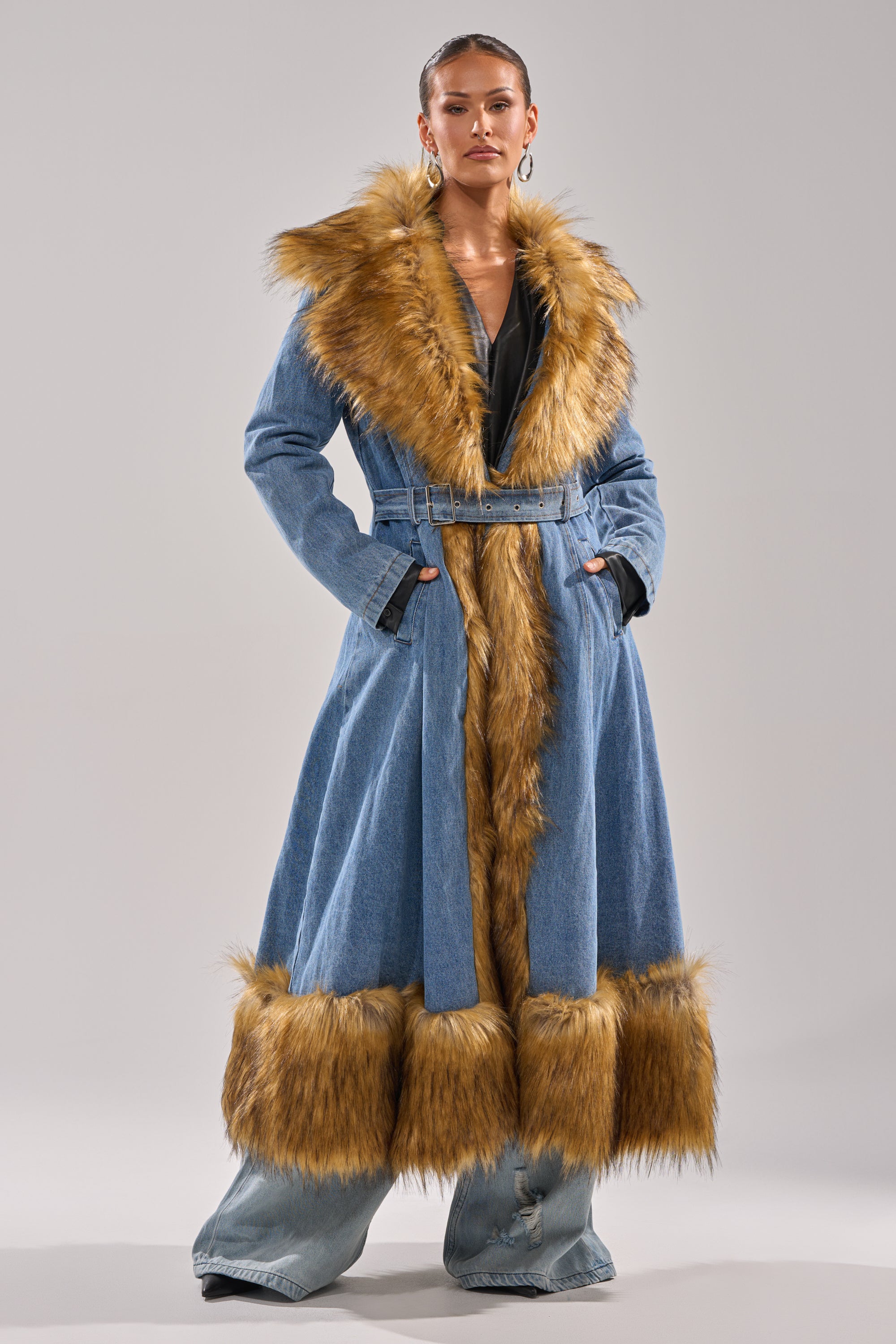 Wearing the VIVIAN FUR TRIM TRENCH IN DENIM with a wide collar, belt, and brown faux fur trim, paired with wide-legged denim pants, this look exudes winter style and confidence against a simple backdrop.