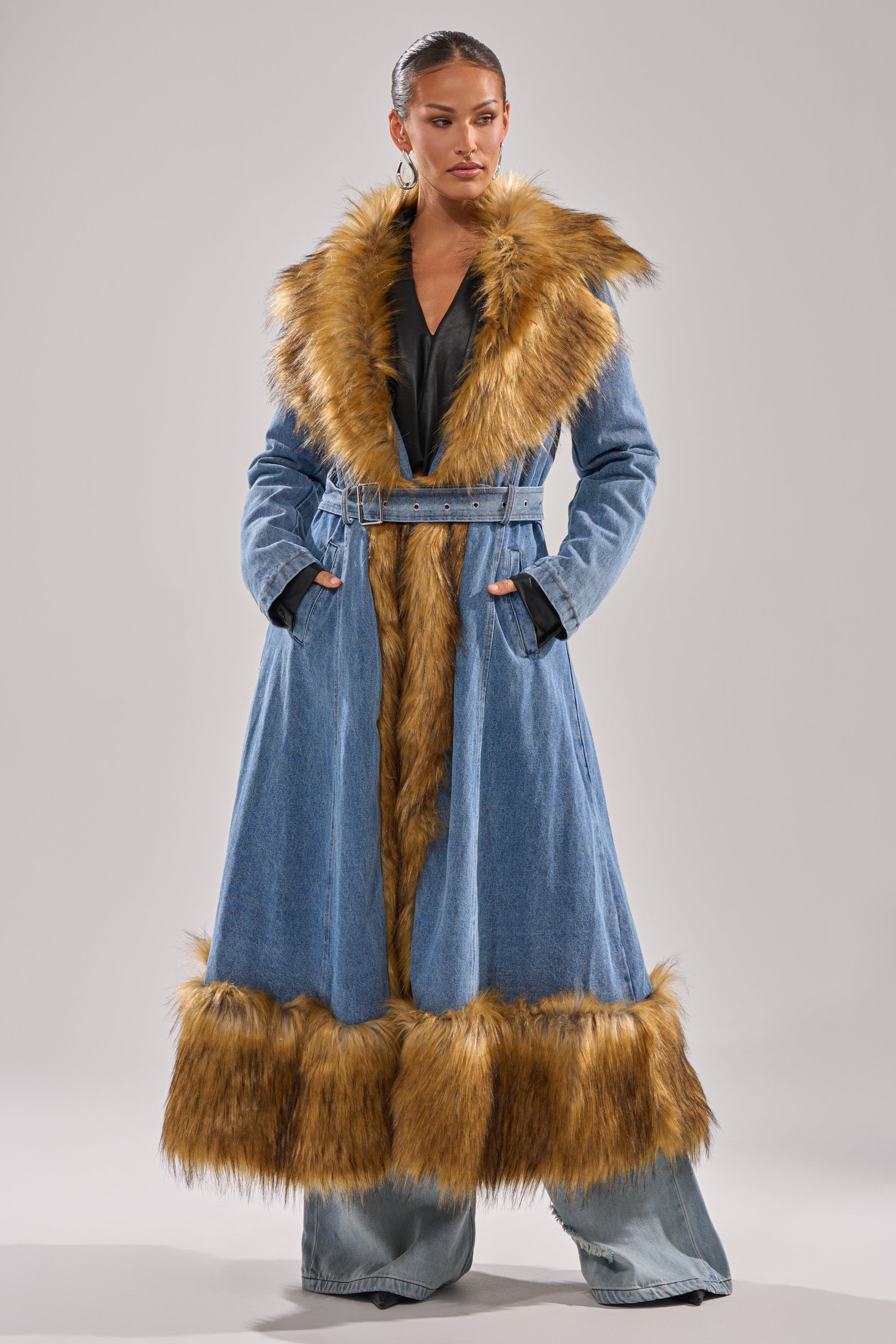 A woman stands confidently in the VIVIAN FUR TRIM TRENCH IN DENIM, featuring wide lapels, golden-brown faux fur trim, a belted waist, and paired with wide-leg denim pants for a bold, modern look.