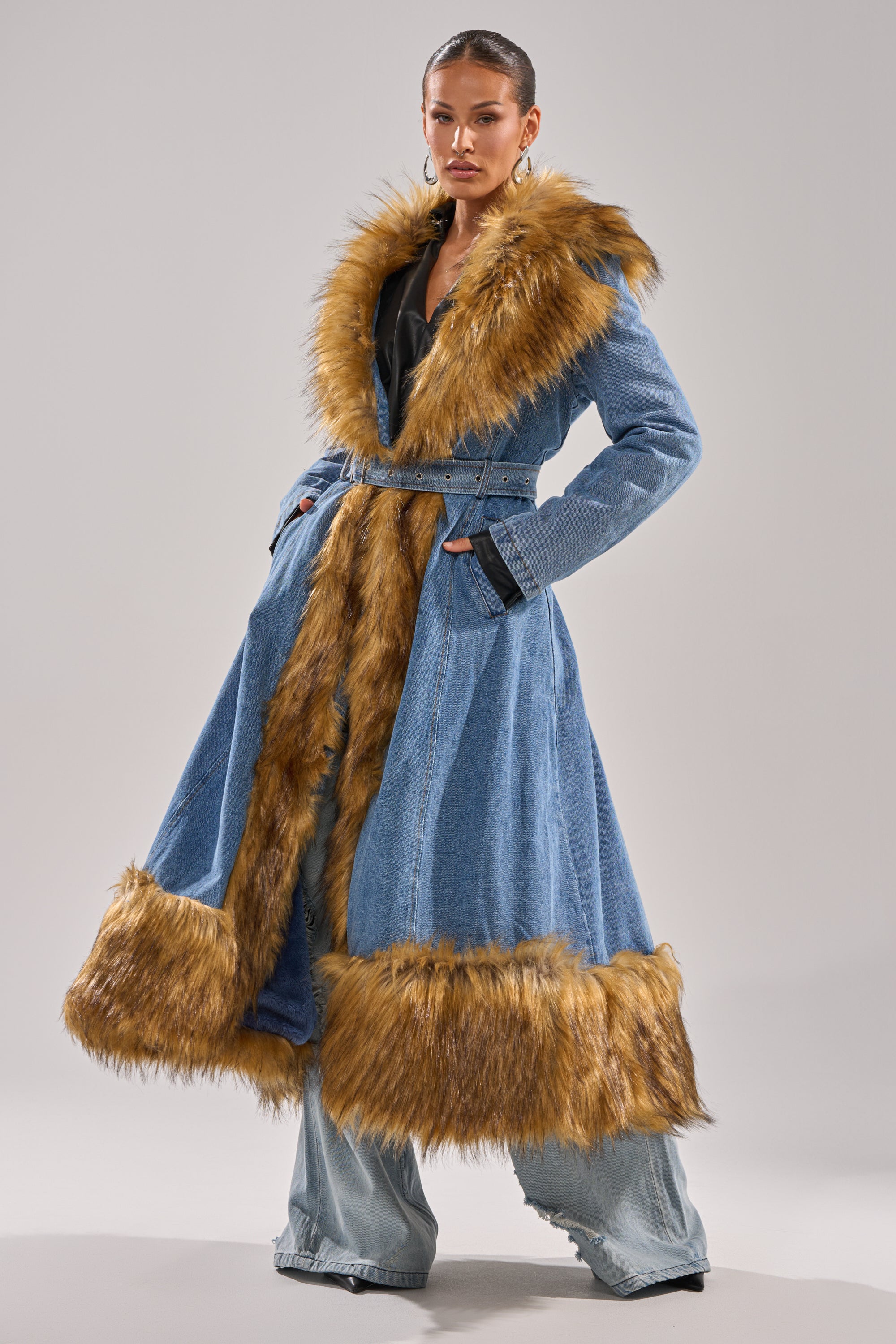 A woman models the VIVIAN FUR TRIM TRENCH IN DENIM, a longline denim coat with brown faux fur on the collar, cuffs, and hem. She pairs it with wide-leg denim pants and hoop earrings against a plain light background.