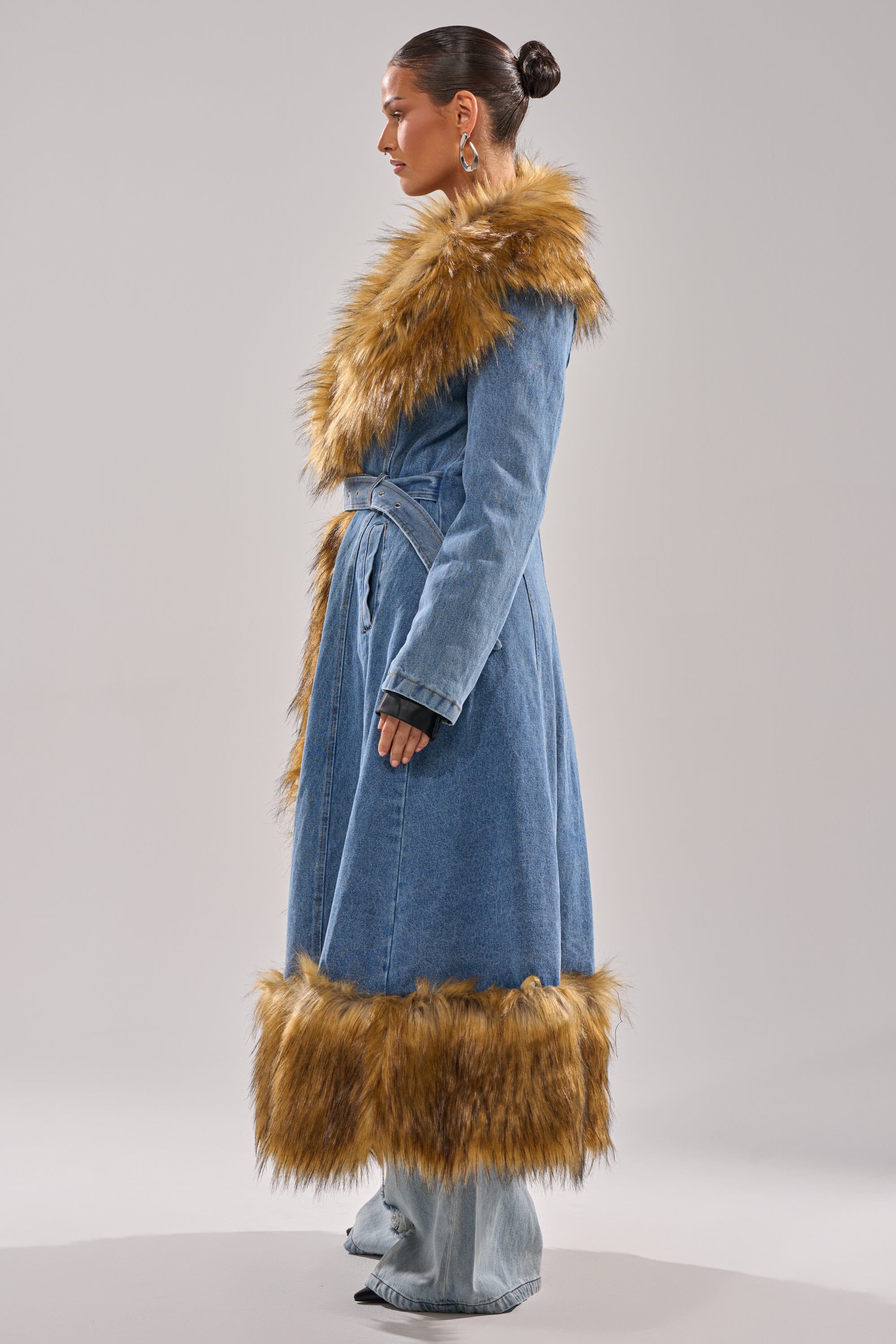 A woman in profile wears the VIVIAN FUR TRIM TRENCH IN DENIM, featuring brown faux fur at the collar, cuffs, and hem. The belted longline coat contrasts with her sleek bun against a plain light background.