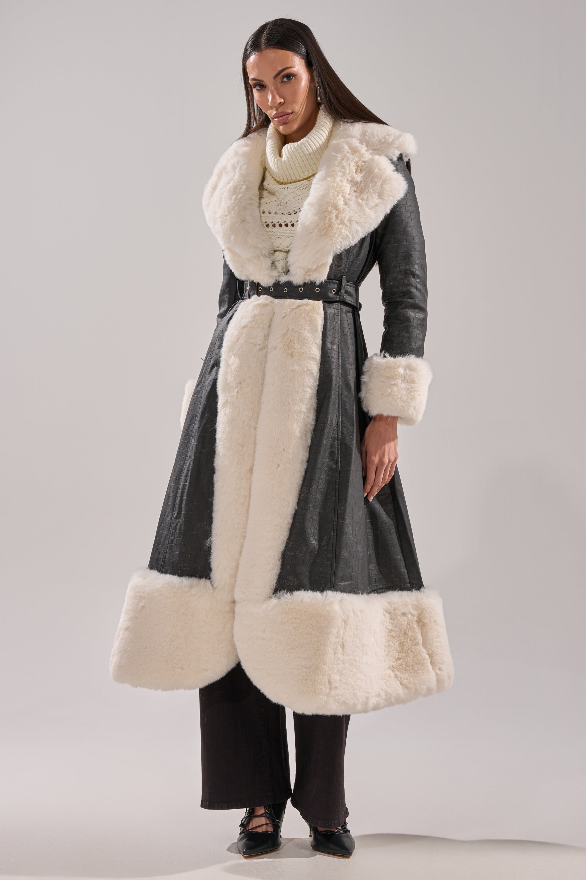 A woman with straight dark hair wears the VIVIAN FUR TRIM TRENCH IN GREY, a long belted faux leather coat featuring wide off-white fur trim on the collar, cuffs, and hem. She pairs it with dark pants and lace-up shoes.