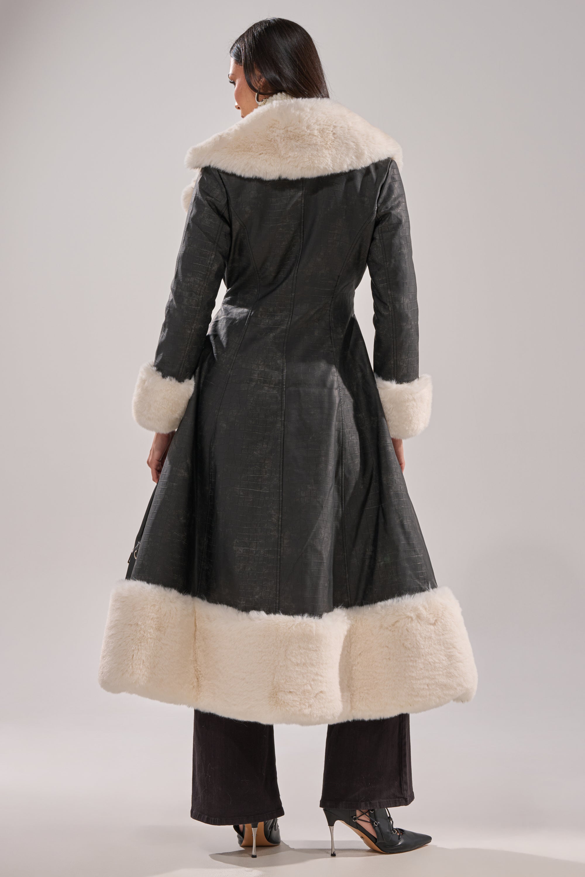 A person faces away, wearing the VIVIAN FUR TRIM TRENCH IN GREY with cream fur on the collar, cuffs, and hem. They style it with dark pants and black heeled shoes against a plain background.