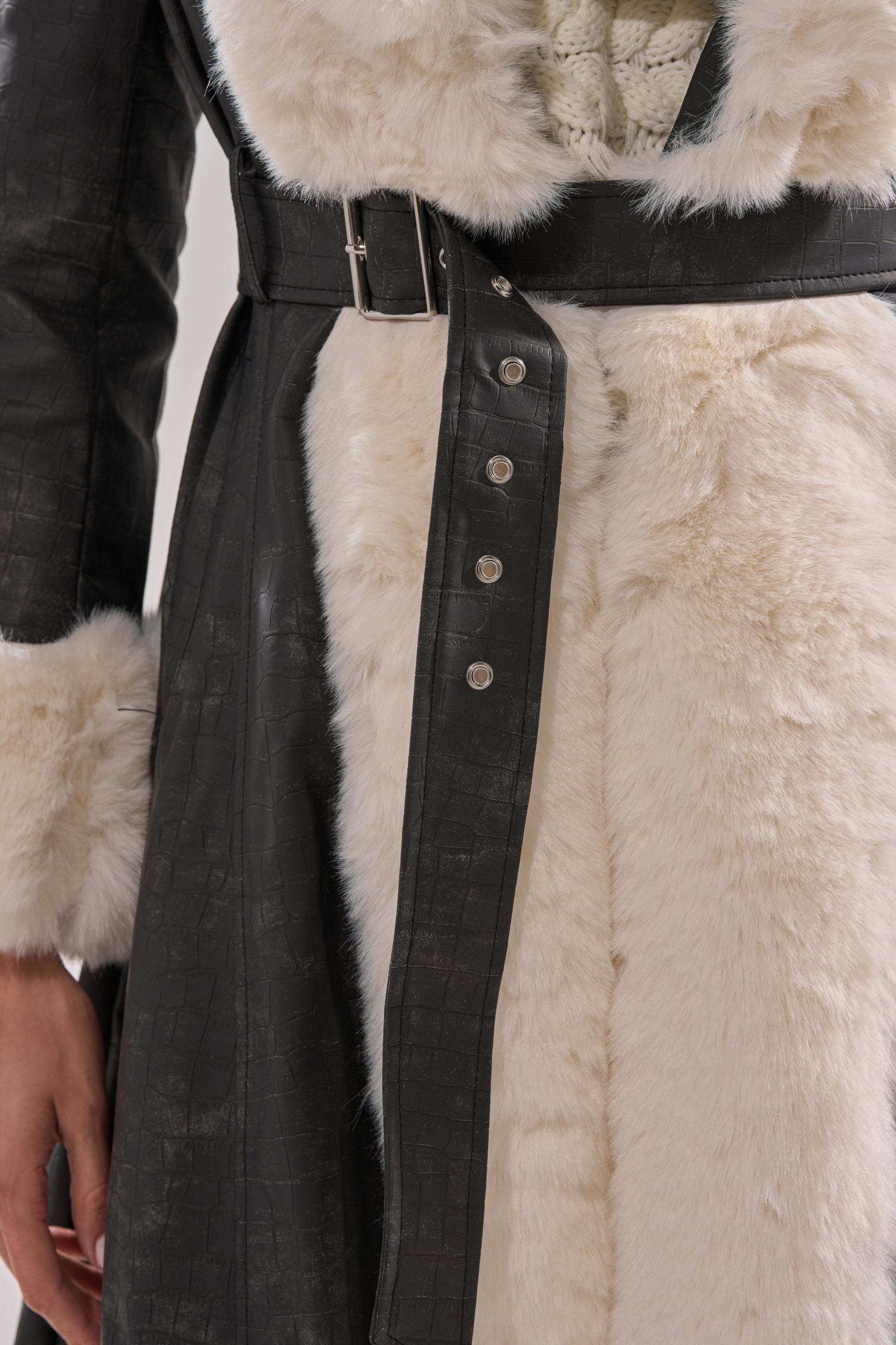 A close-up of a person wearing the VIVIAN FUR TRIM TRENCH IN GREY, featuring faux leather with crocodile texture, white faux fur trim, and a matching belted waist with metallic buttons. Their hand is visible at their side.