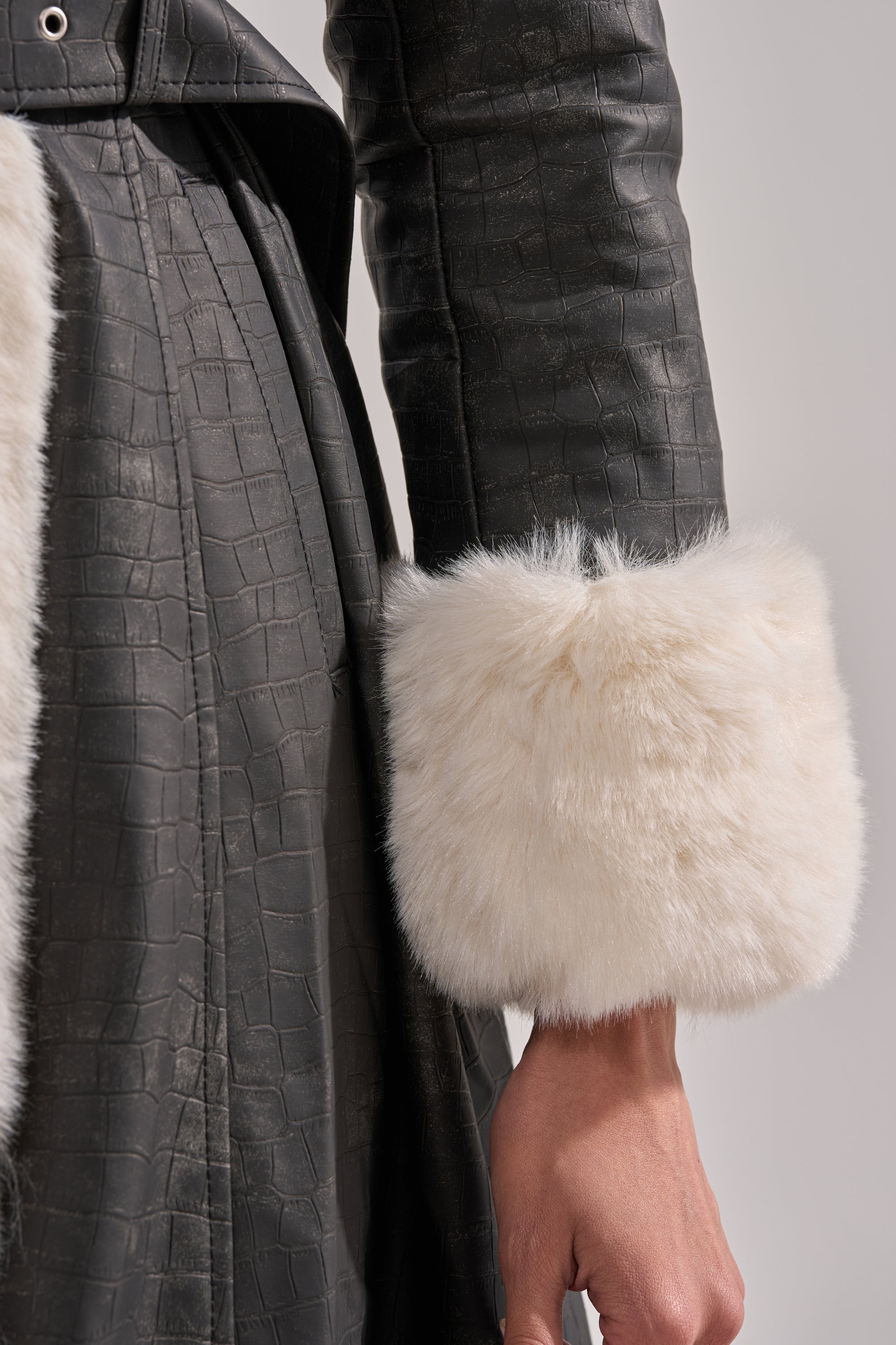 A close-up shows a person wearing the VIVIAN FUR TRIM TRENCH IN GREY, featuring a crocodile print and wide white faux fur cuff, with their hand visible against a neutral background.