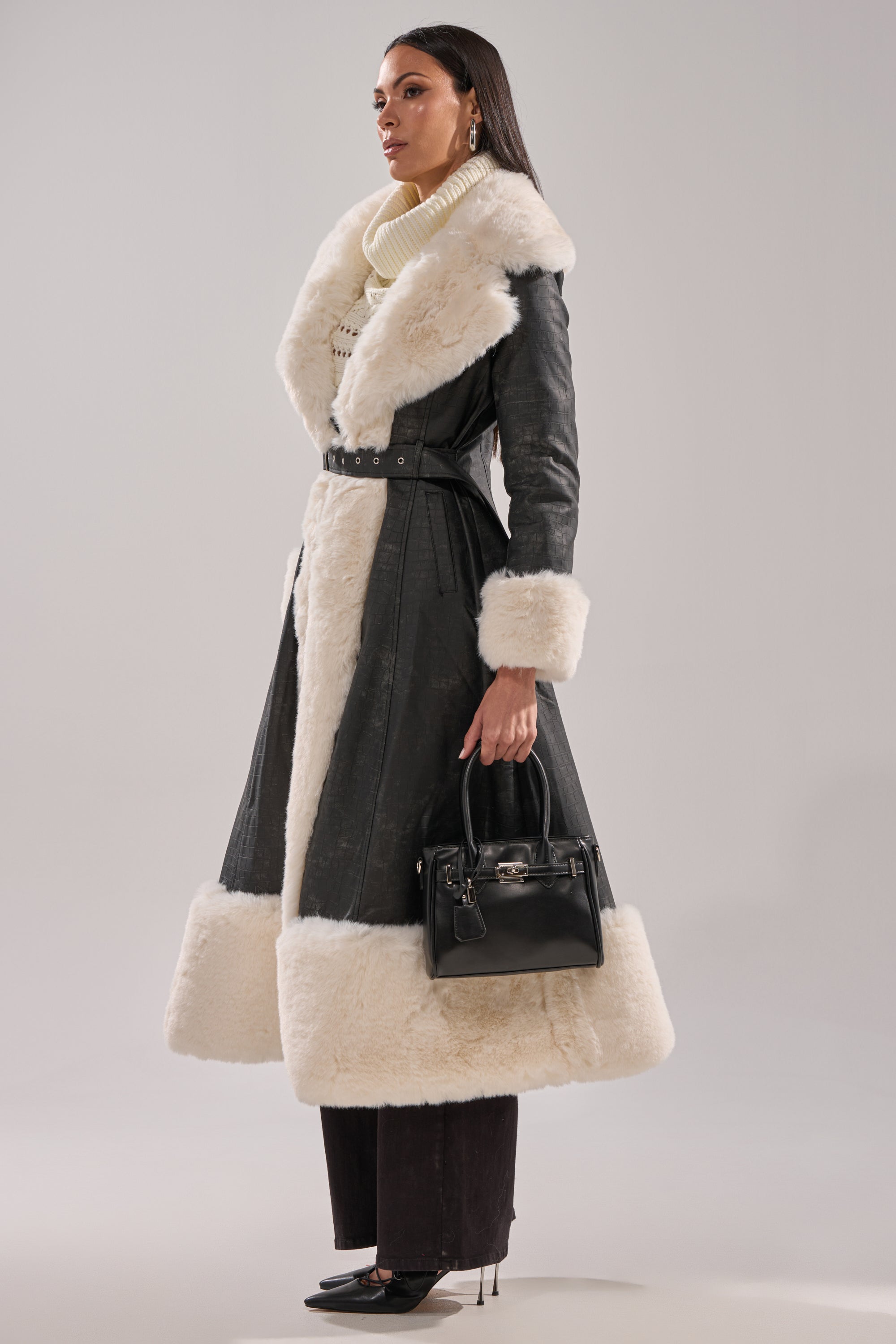 A woman in profile wears the VIVIAN FUR TRIM TRENCH IN GREY with wide white fur, a cream turtleneck, dark pants, and black heels. She carries a black handbag, has long dark hair, and accessorizes with hoop earrings.