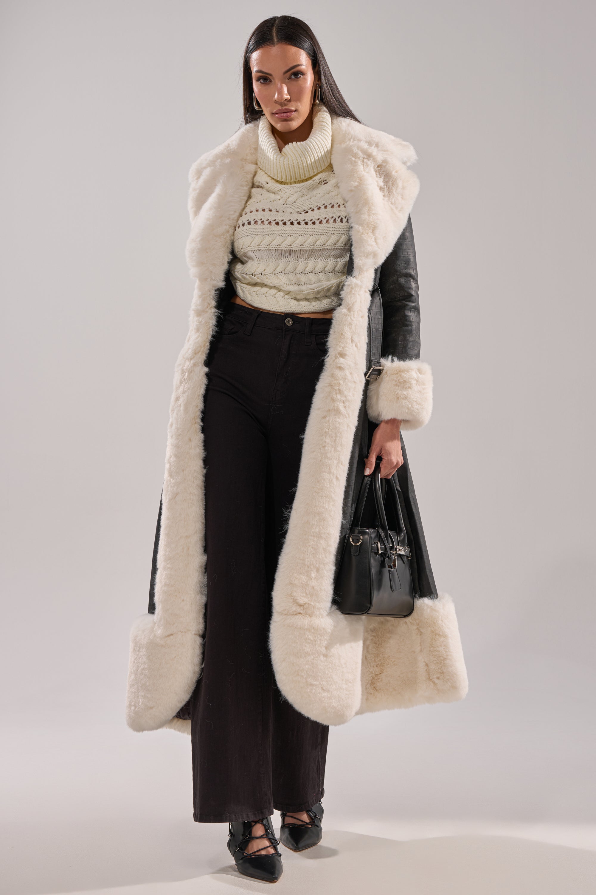 A woman stands confidently in the VIVIAN FUR TRIM TRENCH IN GREY, paired with a cream knit turtleneck, black wide-leg pants, black heels, and a black handbag.