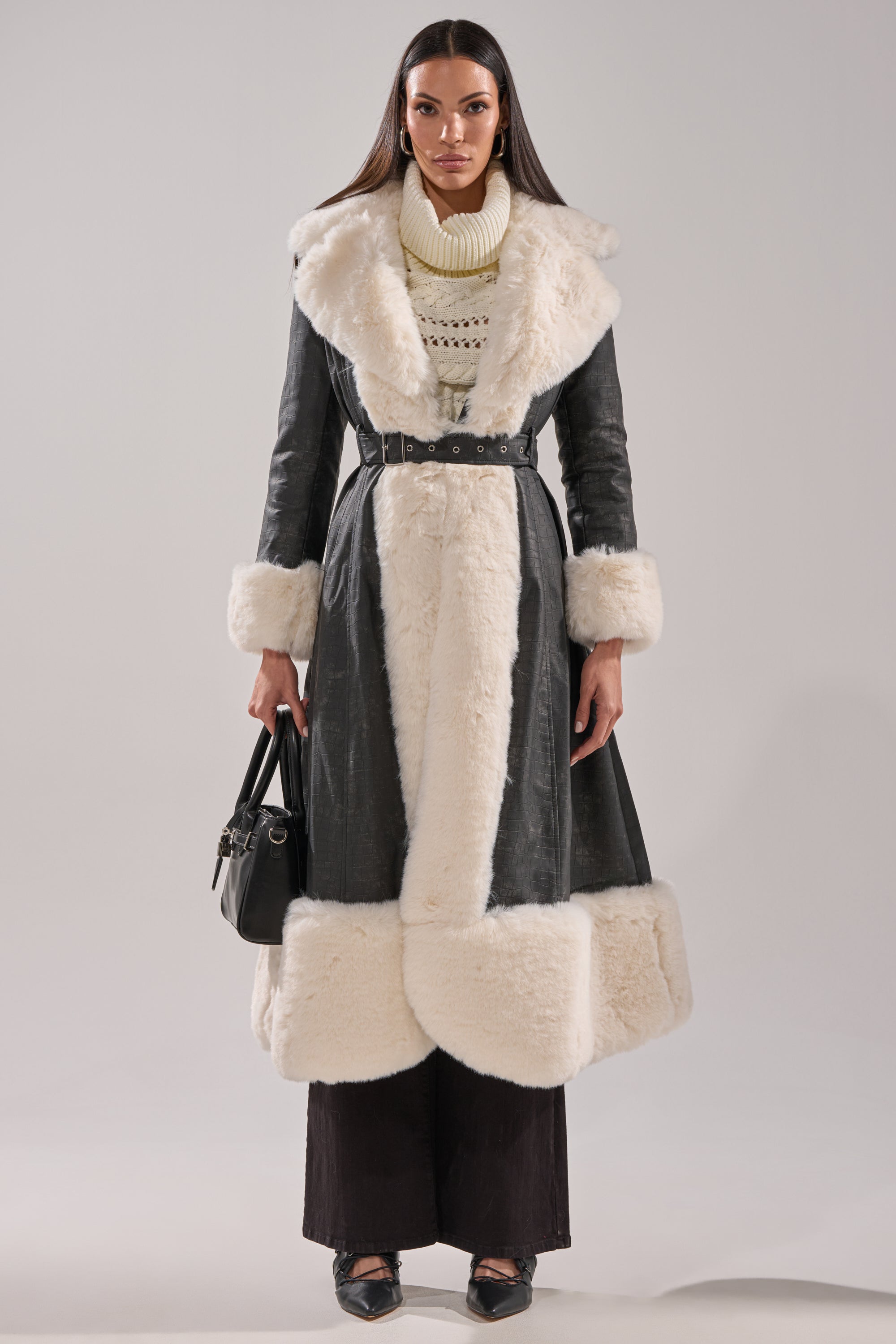 A woman stands facing forward wearing the VIVIAN FUR TRIM TRENCH IN GREY with thick white fur trim, a cream turtleneck sweater, wide-leg dark pants, black shoes, and holding a black handbag.