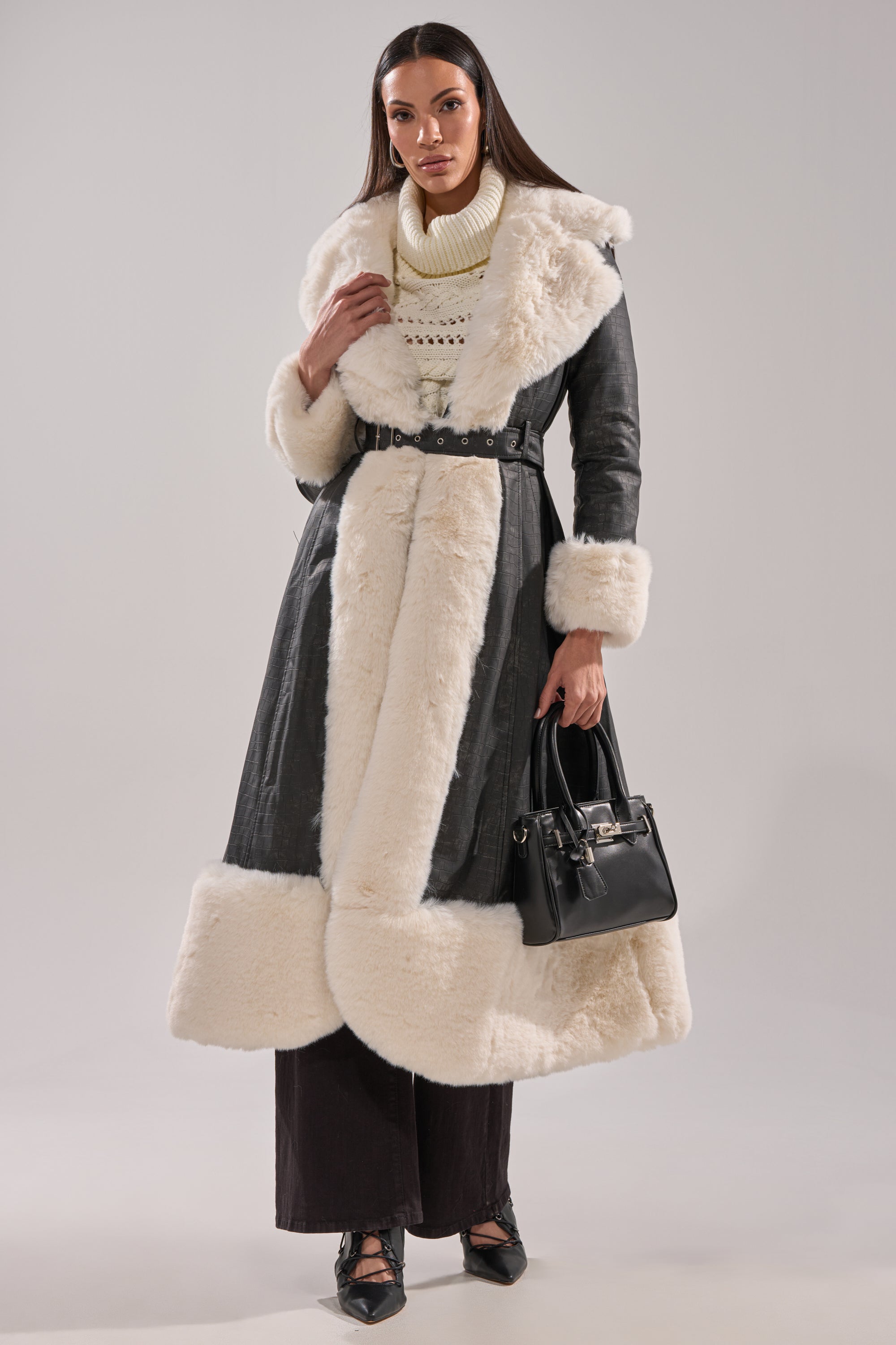 A woman poses against a neutral background wearing the VIVIAN FUR TRIM TRENCH IN GREY with white faux fur accents, a cream knit turtleneck, wide-leg dark pants, black heels, and carrying a black handbag.
