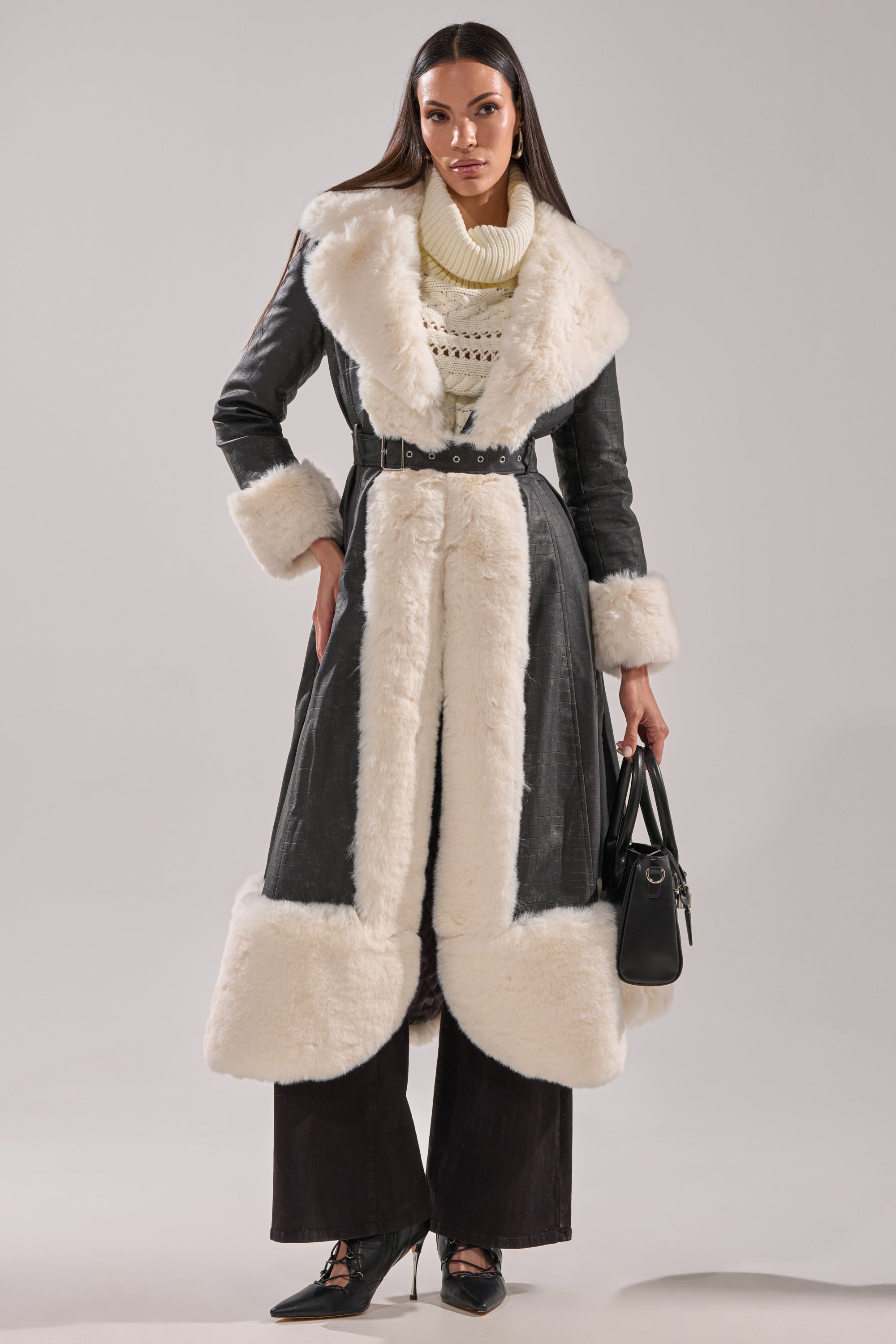 A woman with long straight hair and a confident pose wears the VIVIAN FUR TRIM TRENCH IN GREY, paired with a cream turtleneck sweater, dark pants, black heels, and holds a black handbag.