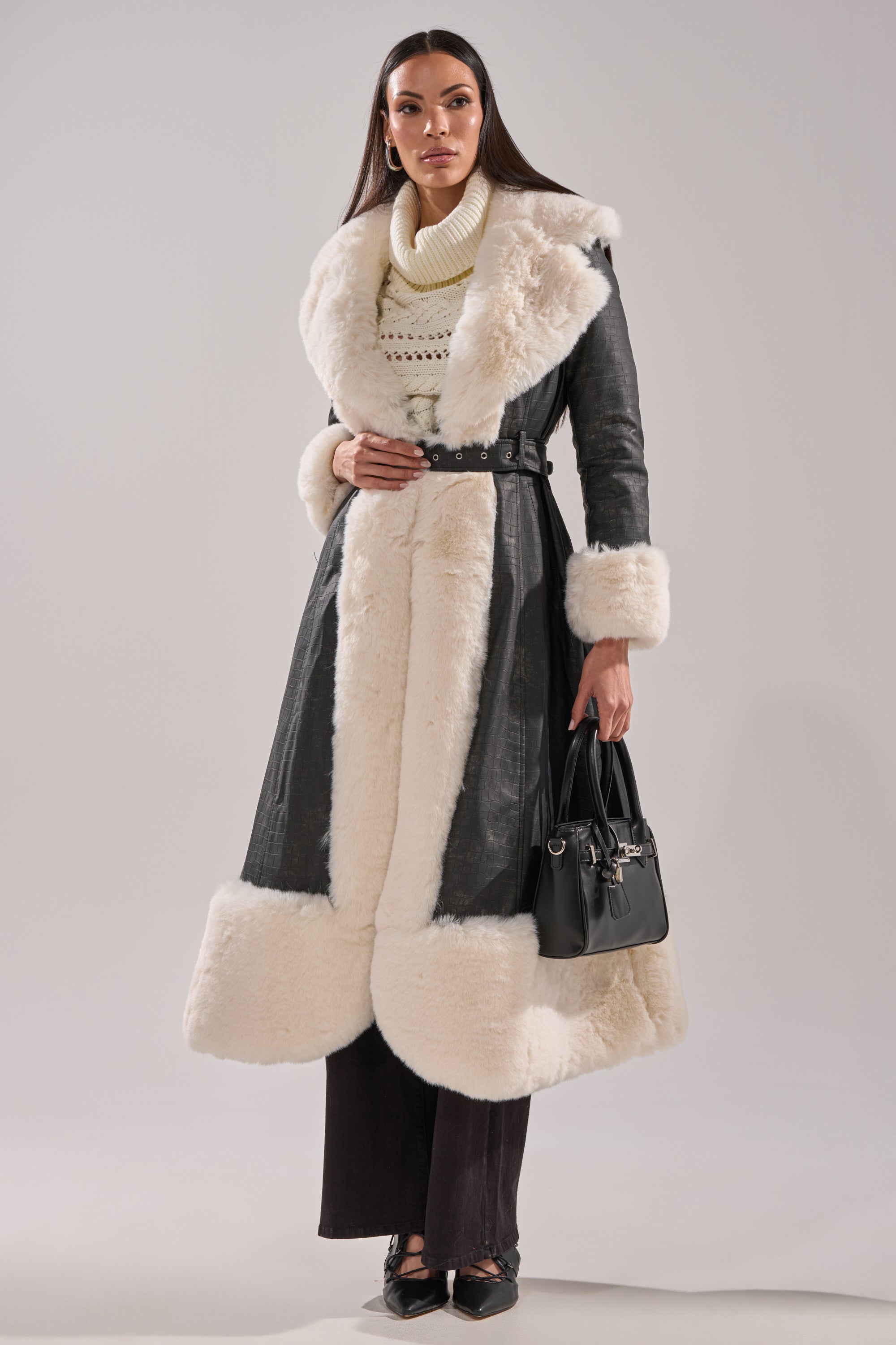 A woman with long straight brown hair stands facing forward, wearing the VIVIAN FUR TRIM TRENCH IN GREY with thick white faux-fur on the collar, cuffs, and hem. She holds a black handbag and wears black pants and shoes.