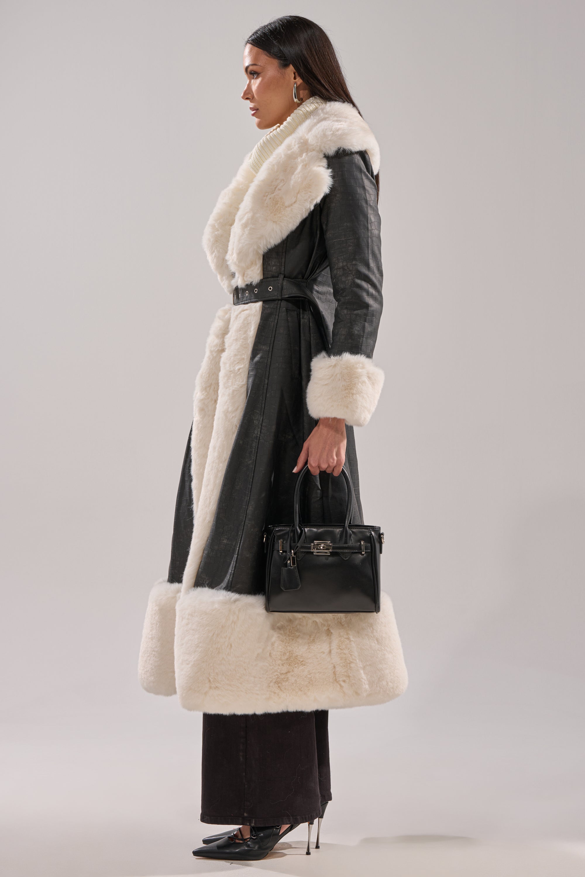 A woman in profile models the VIVIAN FUR TRIM TRENCH IN GREY, paired with a black handbag, dark pants, and black high heels. She has long dark hair and stands against a plain light background.
